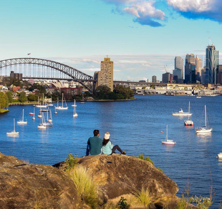 Couple sitting on a rock enjoying a scenic view of Sydney Harbour, with sailboats in the water and the iconic Harbour Bridge in the distance—perfect for a peaceful outing with parents in Sydney.