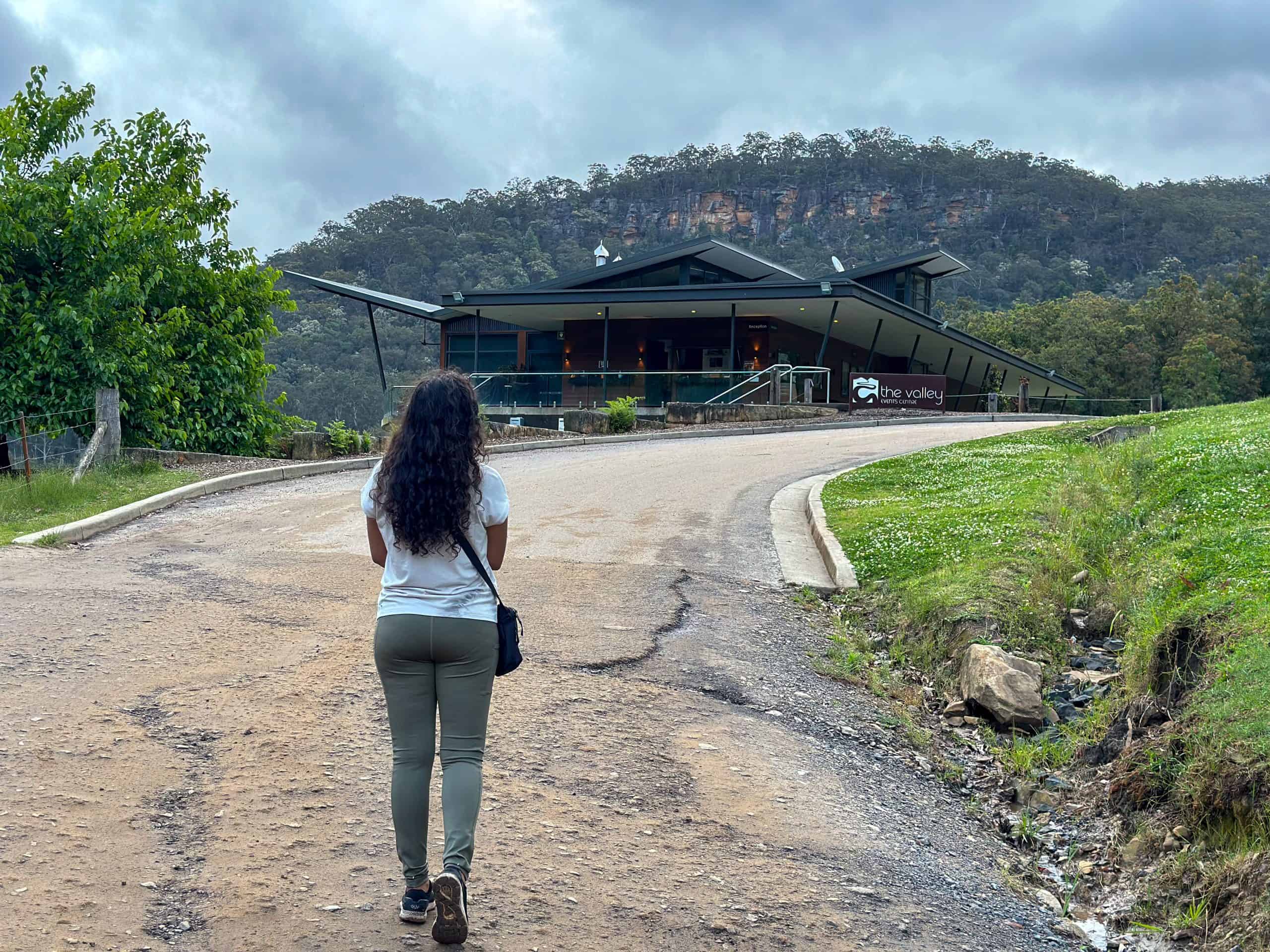A woman walks toward The Valley Wine Cellar, a modern building nestled in lush green hills.