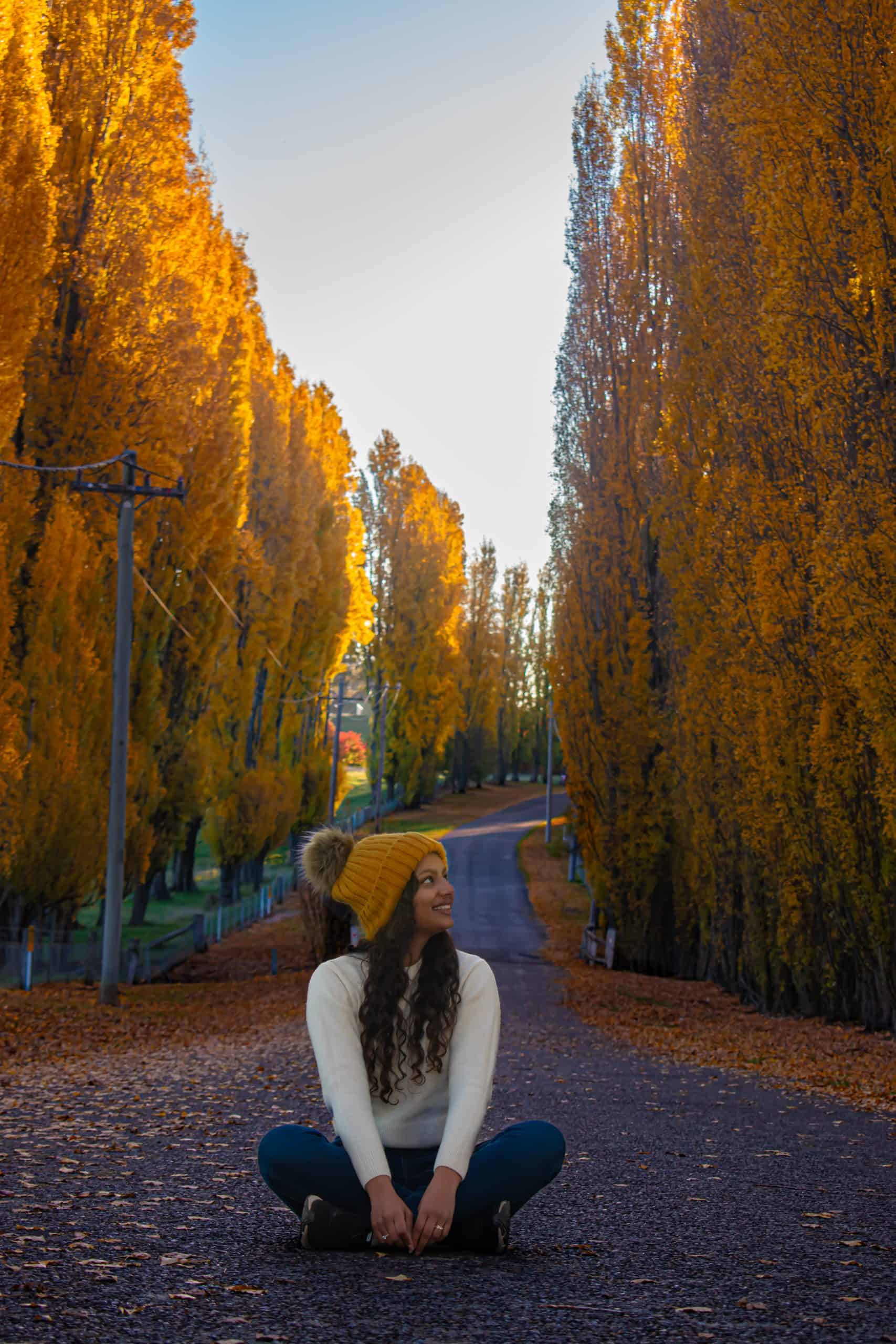 A young woman in a white sweater and mustard beanie sits cross-legged on a quiet road, smiling amidst towering golden poplars in Meadow Flat. The autumn leaves blanket the ground, and the peaceful road stretches into the distance.