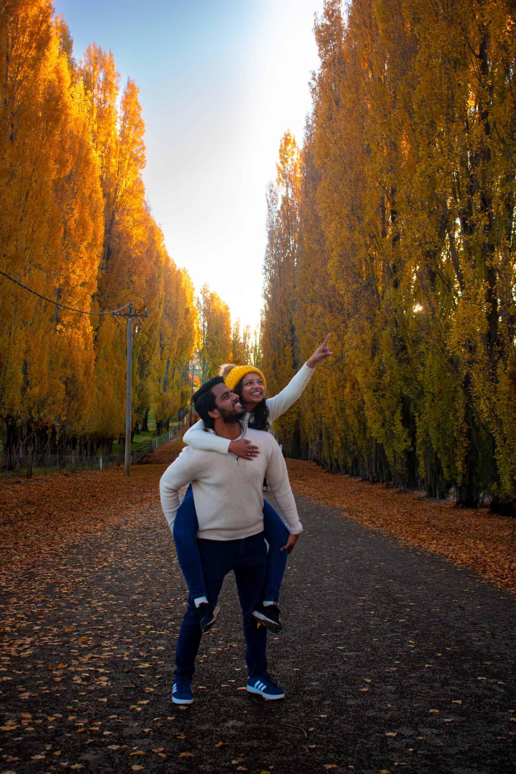  A joyful couple enjoys a piggyback ride under Golden Poplars in Meadow Flat, with the woman playfully pointing towards the sky. The sun casts a warm glow on the autumn leaves, creating a romantic and scenic atmosphere.