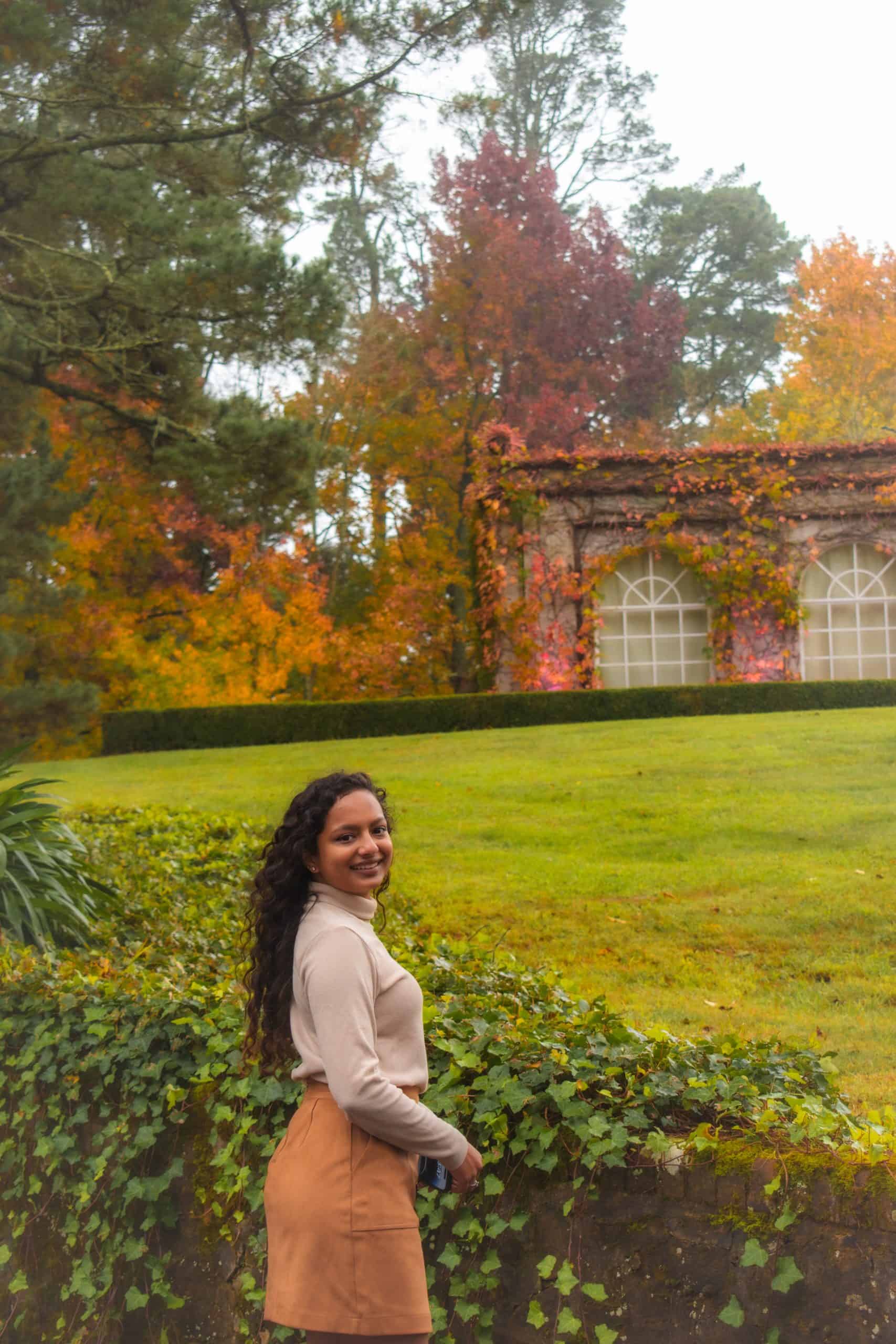 A woman in a beige turtleneck and tan skirt stands by an ivy-covered wall, smiling towards the camera. Behind her, a garden scene with a building covered in autumn leaves adds to the serene ambiance.