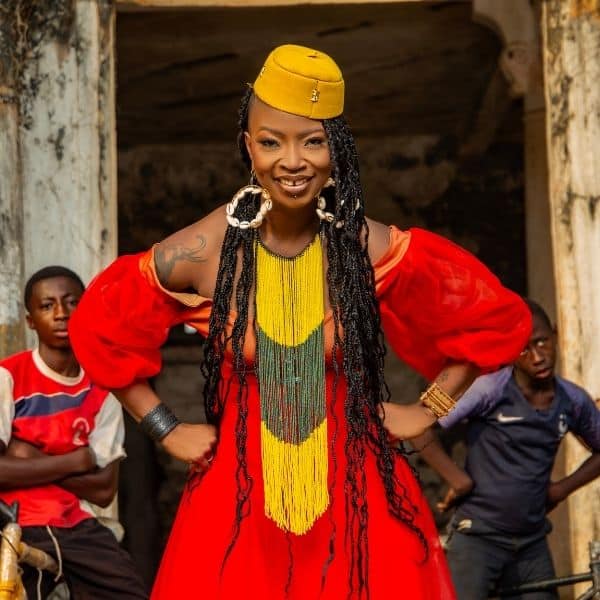 A woman in a vibrant red dress with puffy sleeves and intricate yellow and green beaded jewelry poses confidently. She wears a small yellow hat, large hoop earrings, and long braided hair, with two young boys standing behind her in an outdoor setting