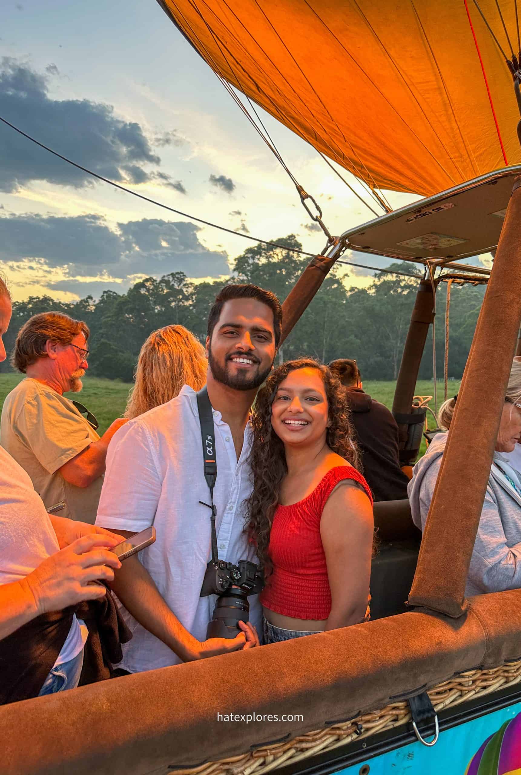 A man in a white shirt, with a camera around his neck, is posing with a girl in a red top in a hot air balloon basket.