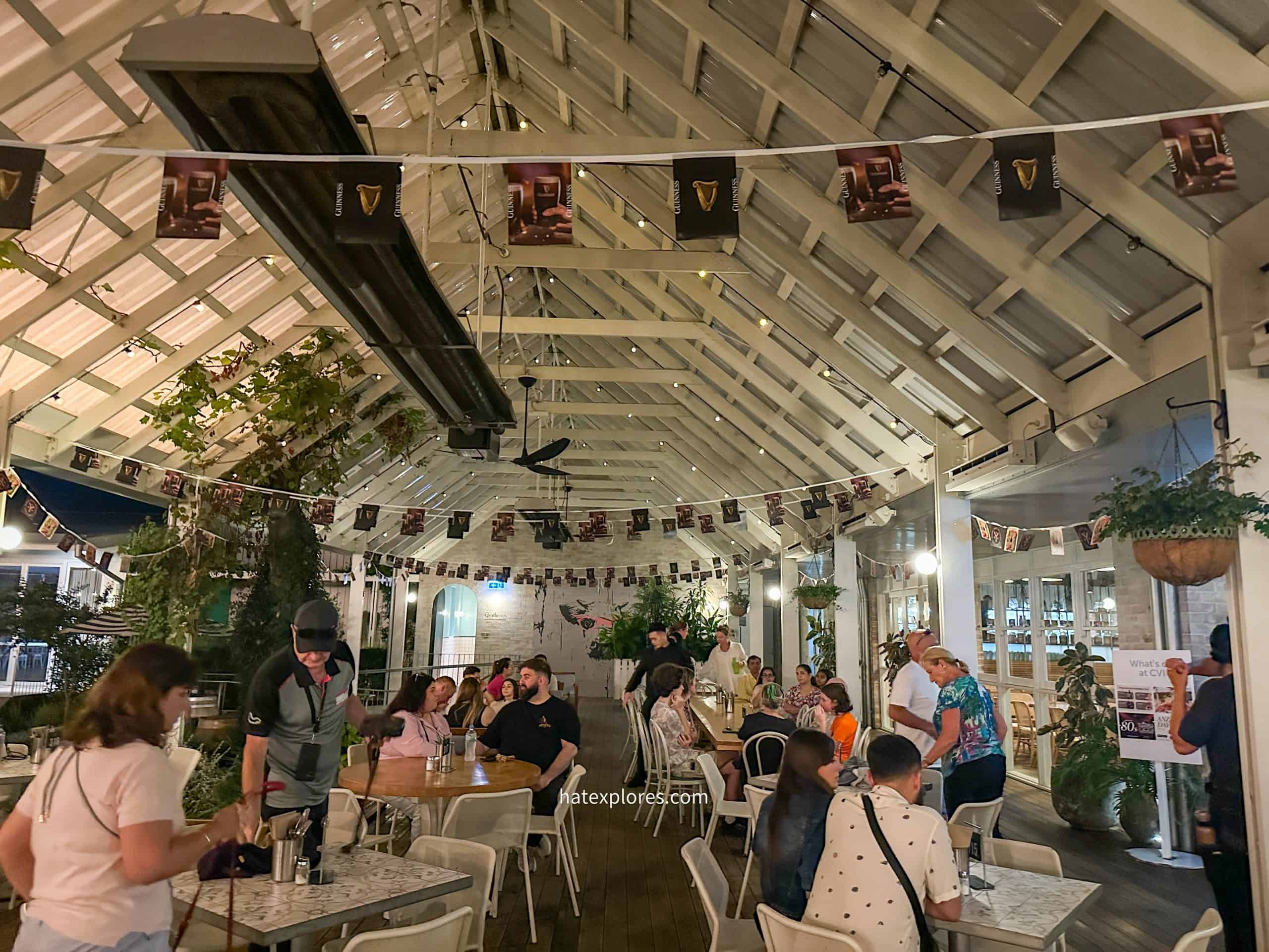 A lively indoor-outdoor restaurant or bar with a high, white wooden ceiling adorned with Guinness-branded bunting. Patrons are seated at white tables, engaged in conversation, while some people stand or move around. Hanging plants, warm lighting, and a mix of modern and rustic decor create a cozy atmosphere. Scenes before a hot air balloon ride in Sydney! 