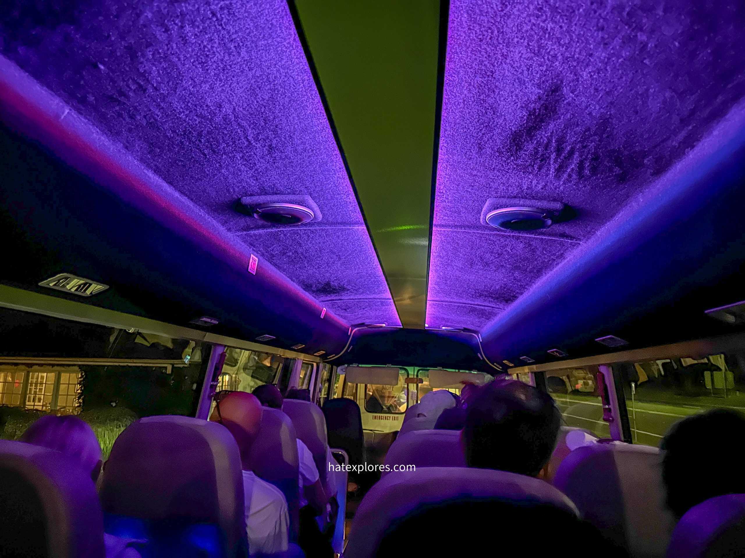 The interior of a bus at night, illuminated by vibrant purple and blue LED lighting along the ceiling. Passengers sit in rows of cushioned seats, facing forward, with some silhouettes visible against the dimly lit windows. The driver’s area is visible at the front, showing reflections of streetlights and buildings outside.