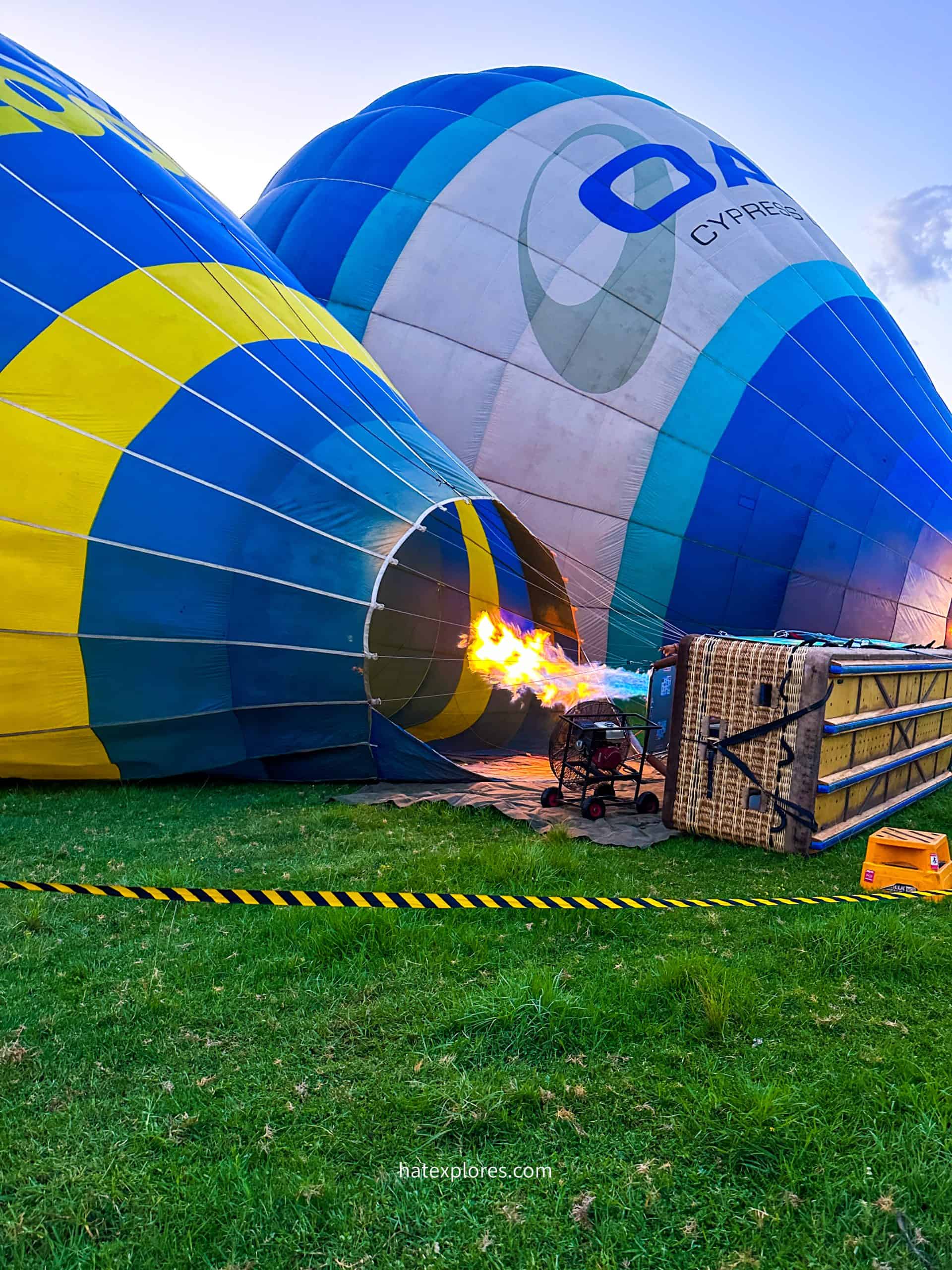 A hot air balloon ride in Sydney being inflated with a bright flame shooting into its blue and yellow envelope from a burner. The partially inflated balloon lies on a grassy field, with another blue and white balloon in the background. A wicker basket rests on its side, and a caution tape barrier runs across the foreground.