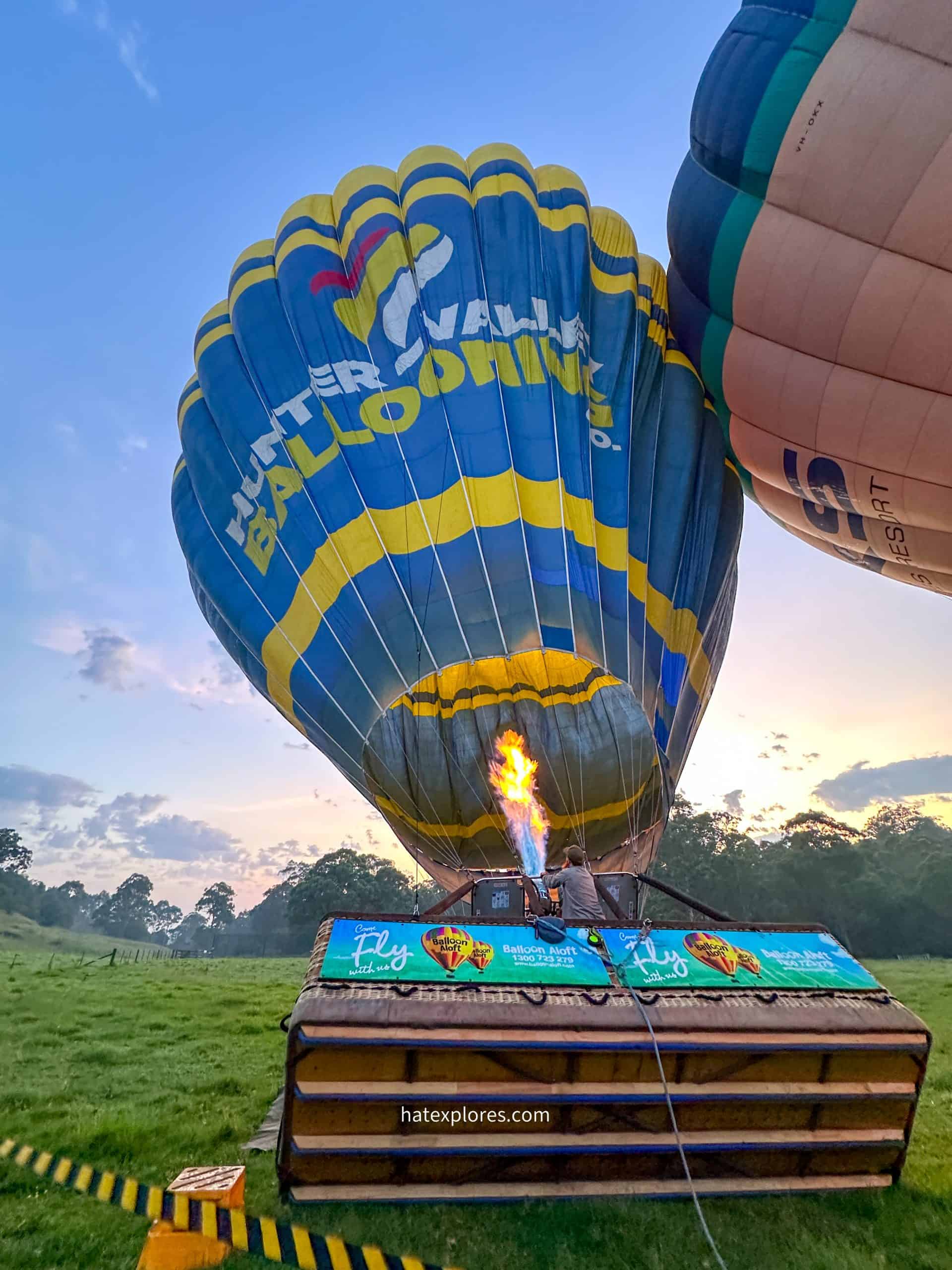 A hot air balloon is being inflated as a powerful flame shoots into its blue and yellow envelope. The wicker basket lies on its side nearby, and another large blue and white balloon with "OAK CYPRESS" printed on it is partially inflated in the background. A black and yellow caution tape runs across the grassy field in the foreground