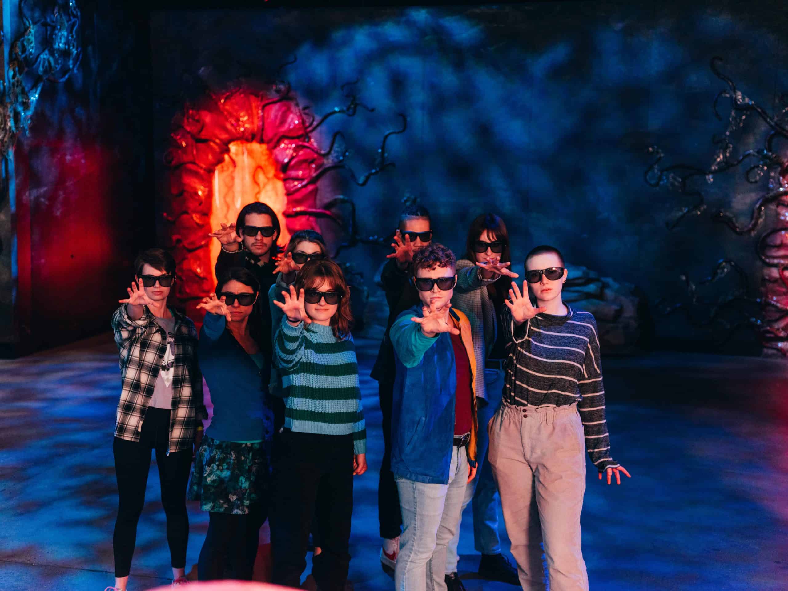 A group of people wearing 3D glasses strike a synchronized hand gesture in a dimly lit room. A glowing red portal-like structure and eerie blue lighting create an immersive, sci-fi atmosphere.