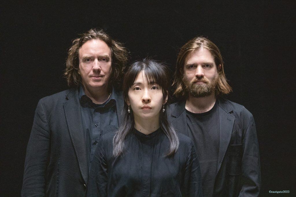 Three musicians dressed in black stand against a dark background. The central figure is a young woman with shoulder-length dark hair and silver earrings, flanked by two men with serious expressions, one with wavy brown hair and the other with a beard.