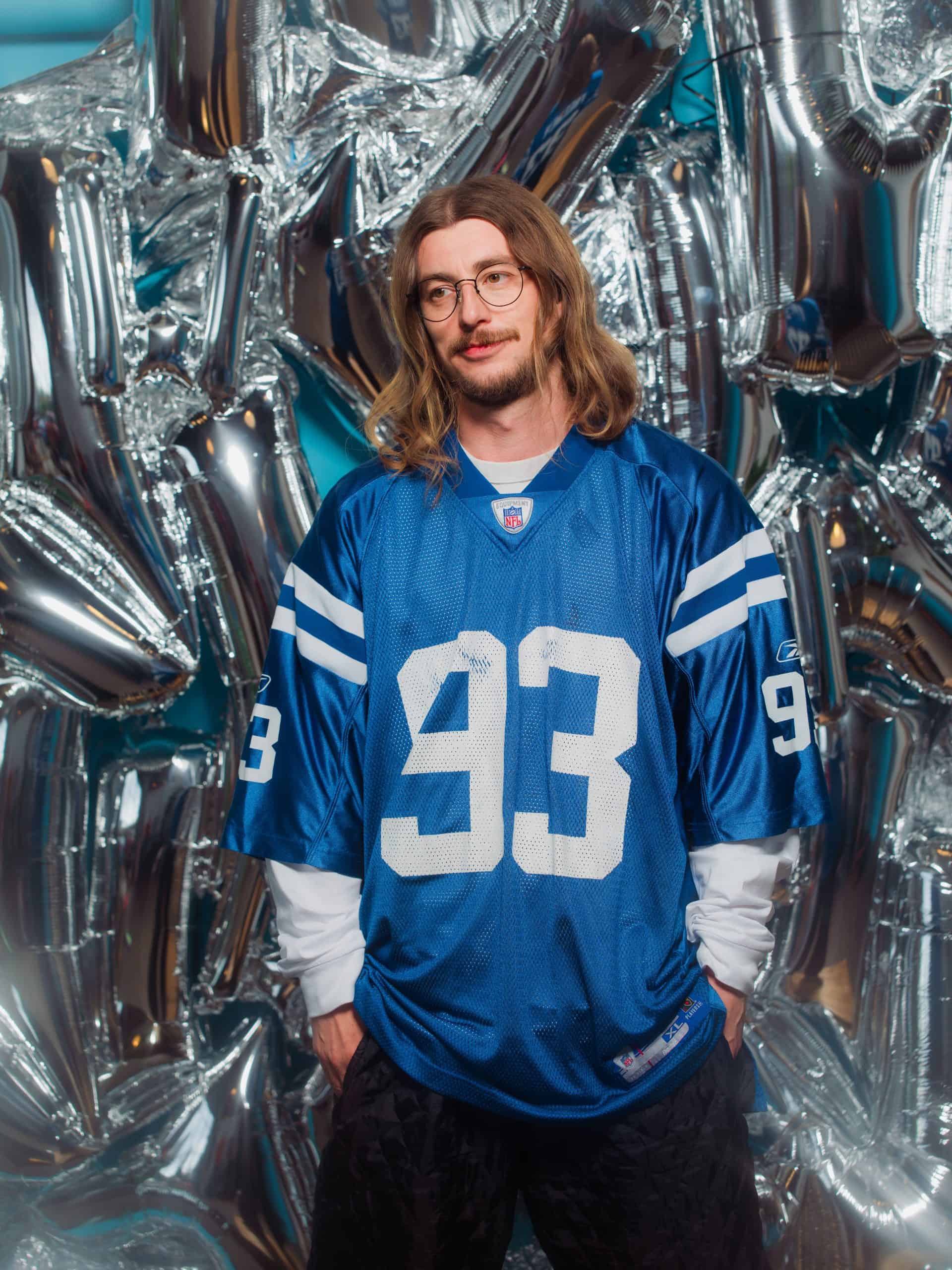 A man with shoulder-length wavy hair, glasses, and facial hair wears a blue Indianapolis Colts jersey with the number 93. He stands in front of silver balloon decorations, looking relaxed and slightly smiling.