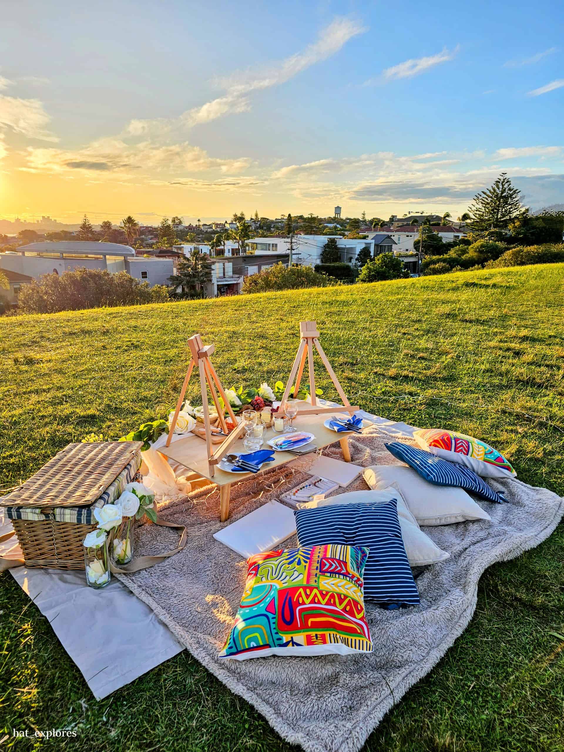 A cozy picnic setup with bright cushions, a wicker basket, and a small wooden table overlooks the Sydney skyline from a sunny hilltop at Dudley Page Reserve—an inviting scene that highlights one of the best free things to do in Sydney.
