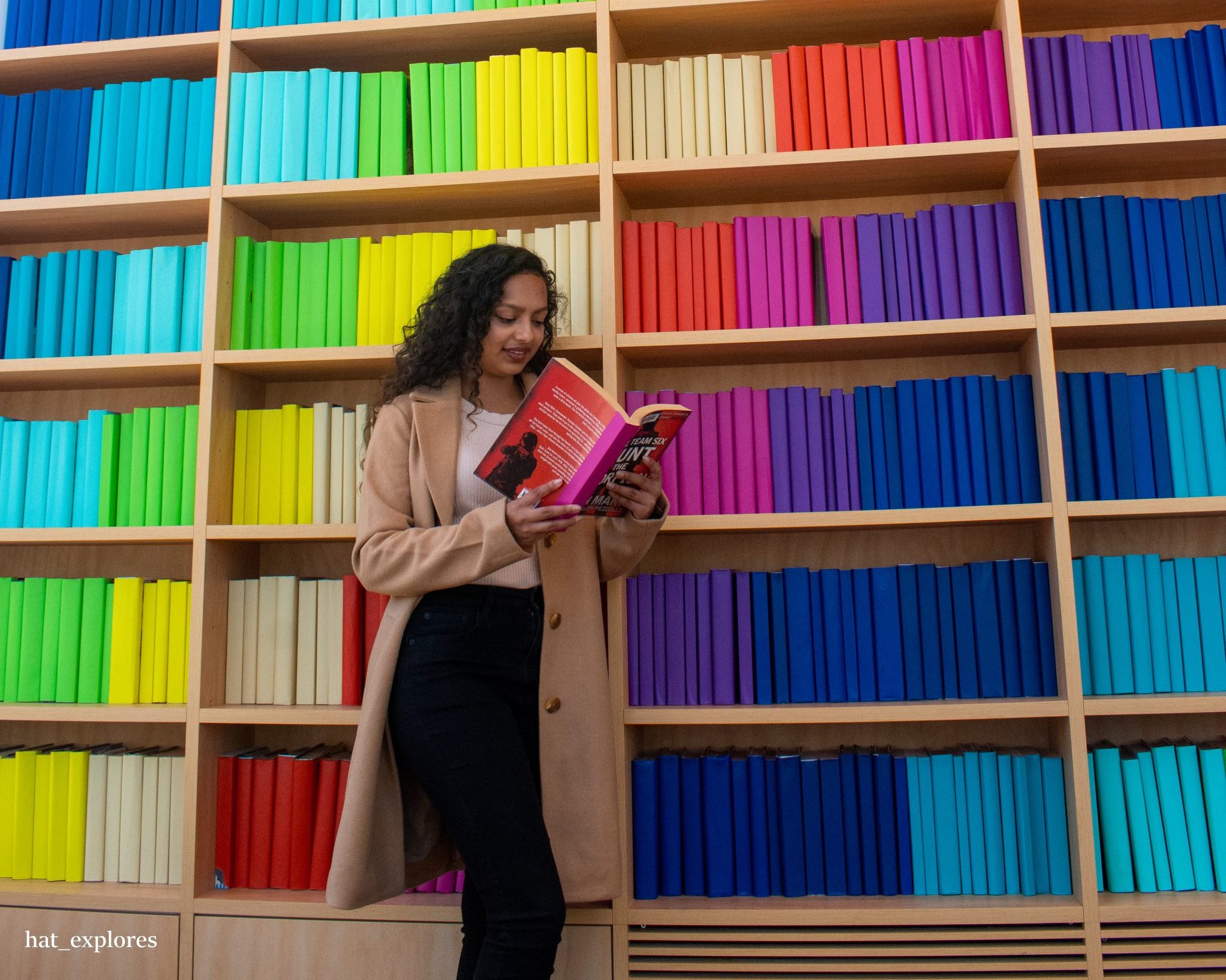 A girl is reading a book taken from a shelf with colourful ones aligned beautifully in a pattern.