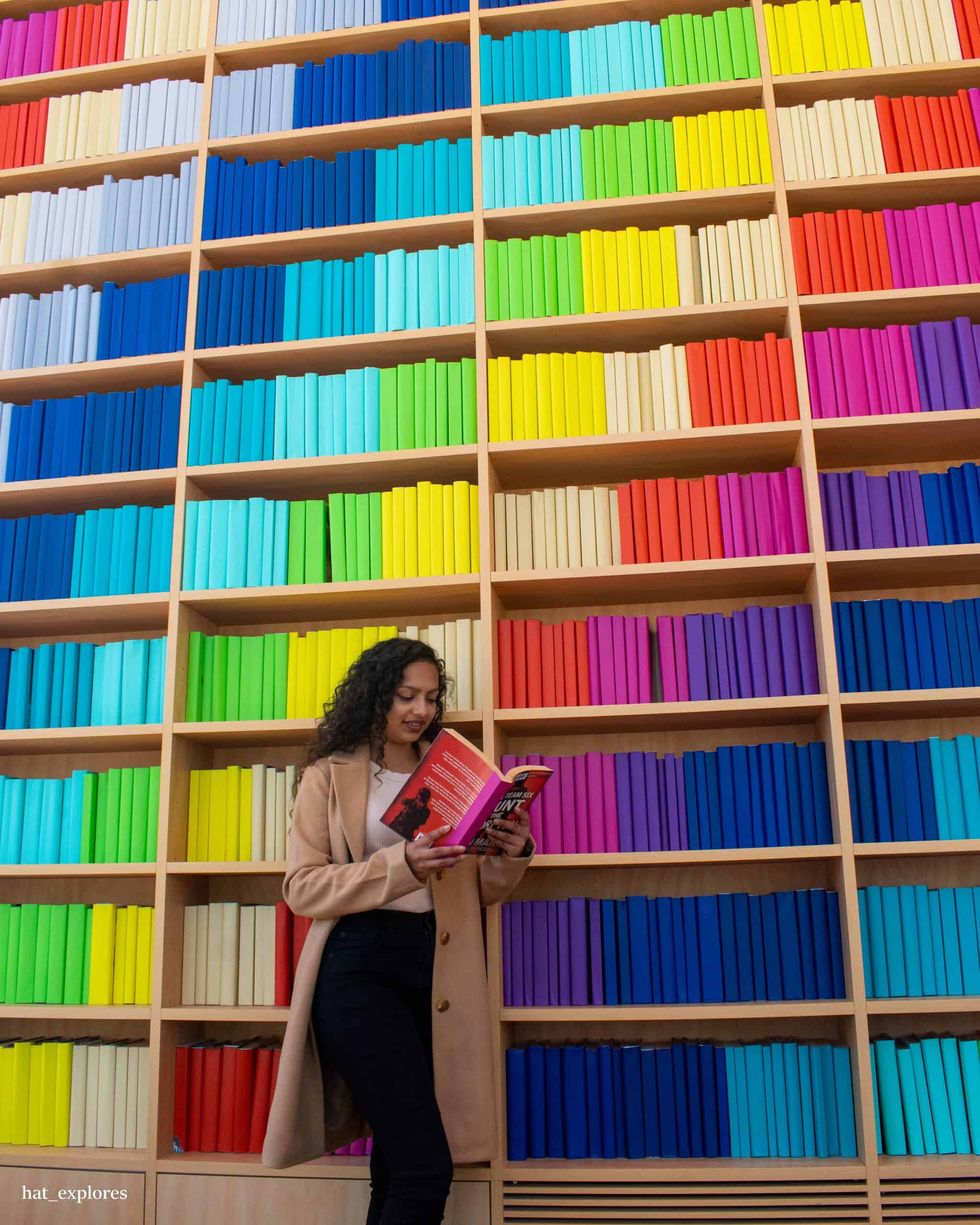 A girl reading a book in Green Square Library, were the books behind her are aligned in a colourful pattern.