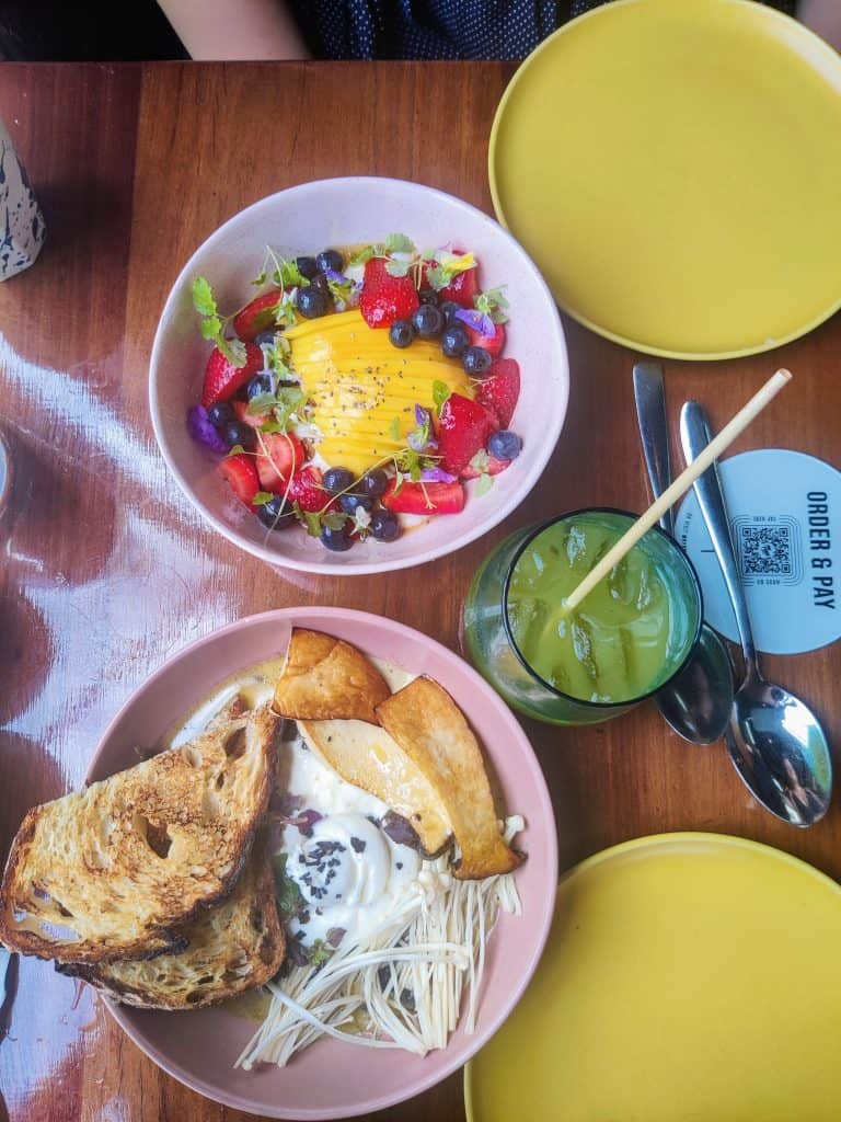 A table full of fresh food bowls with berries, strawberries, and a slice of mango cut into vertical strips in one and a glass of matcha coffee to compliment it.