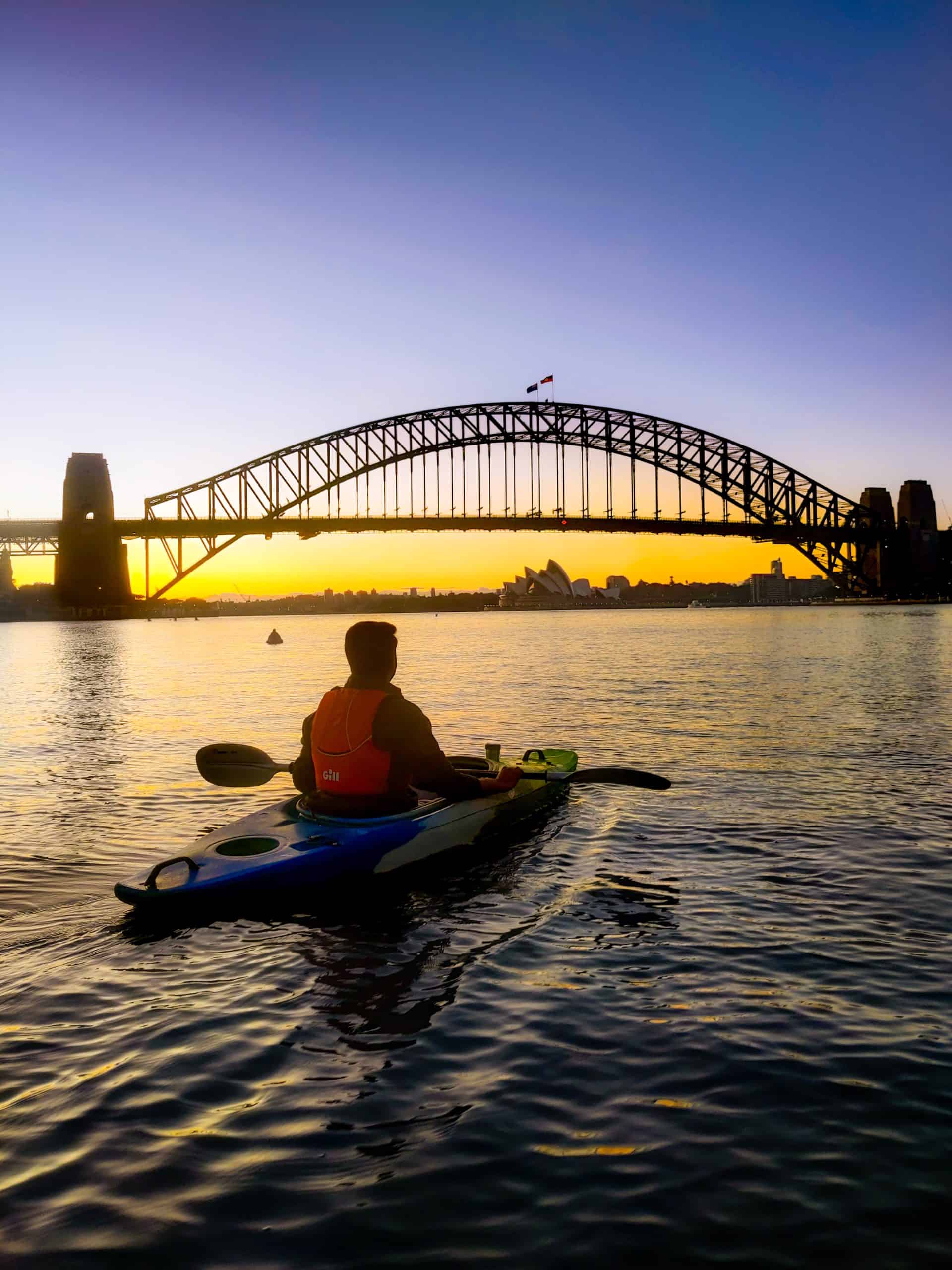 Person kayaking at dawn in Sydney Harbour with the Harbour Bridge and Opera House in the distance, offering a unique morning activity for a one day Sydney itinerary.