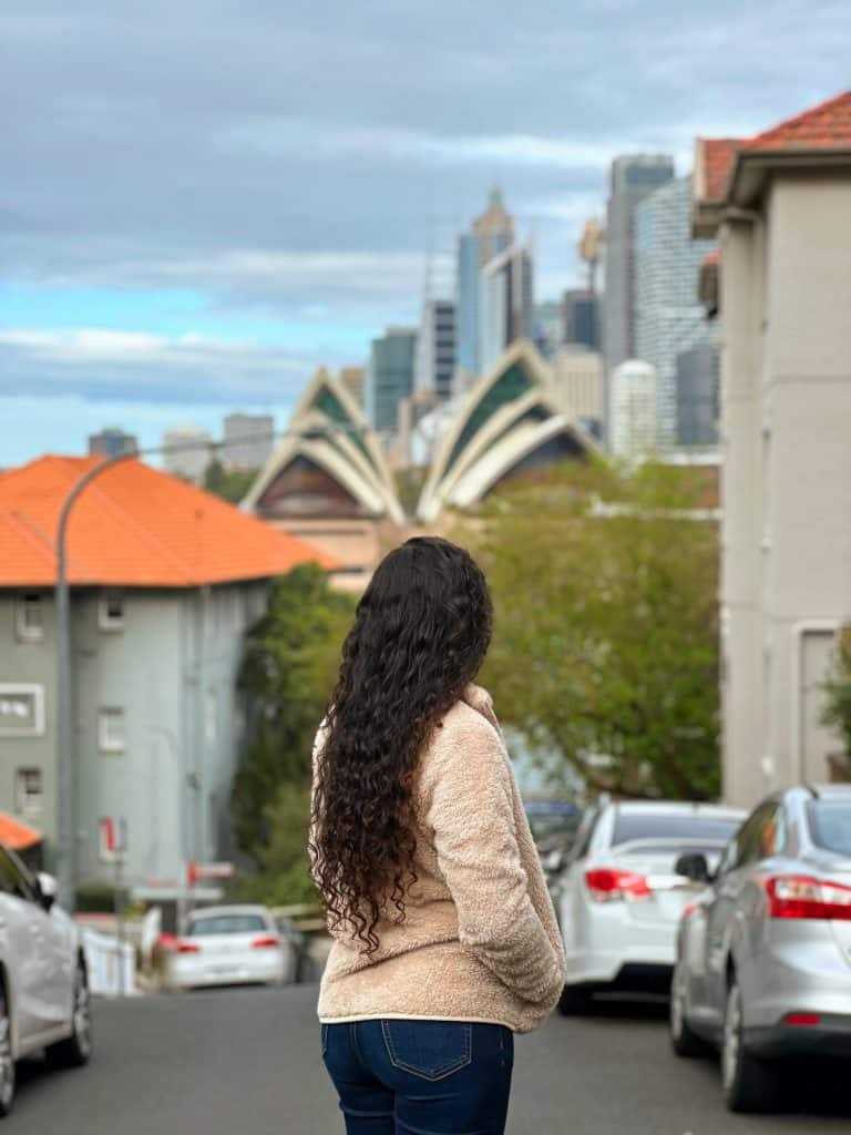  A woman with long curly hair stands on a quiet street in Kirribilli, gazing toward the iconic Sydney Opera House and city skyline—one of the best viewpoints and things to do in Kirribilli.