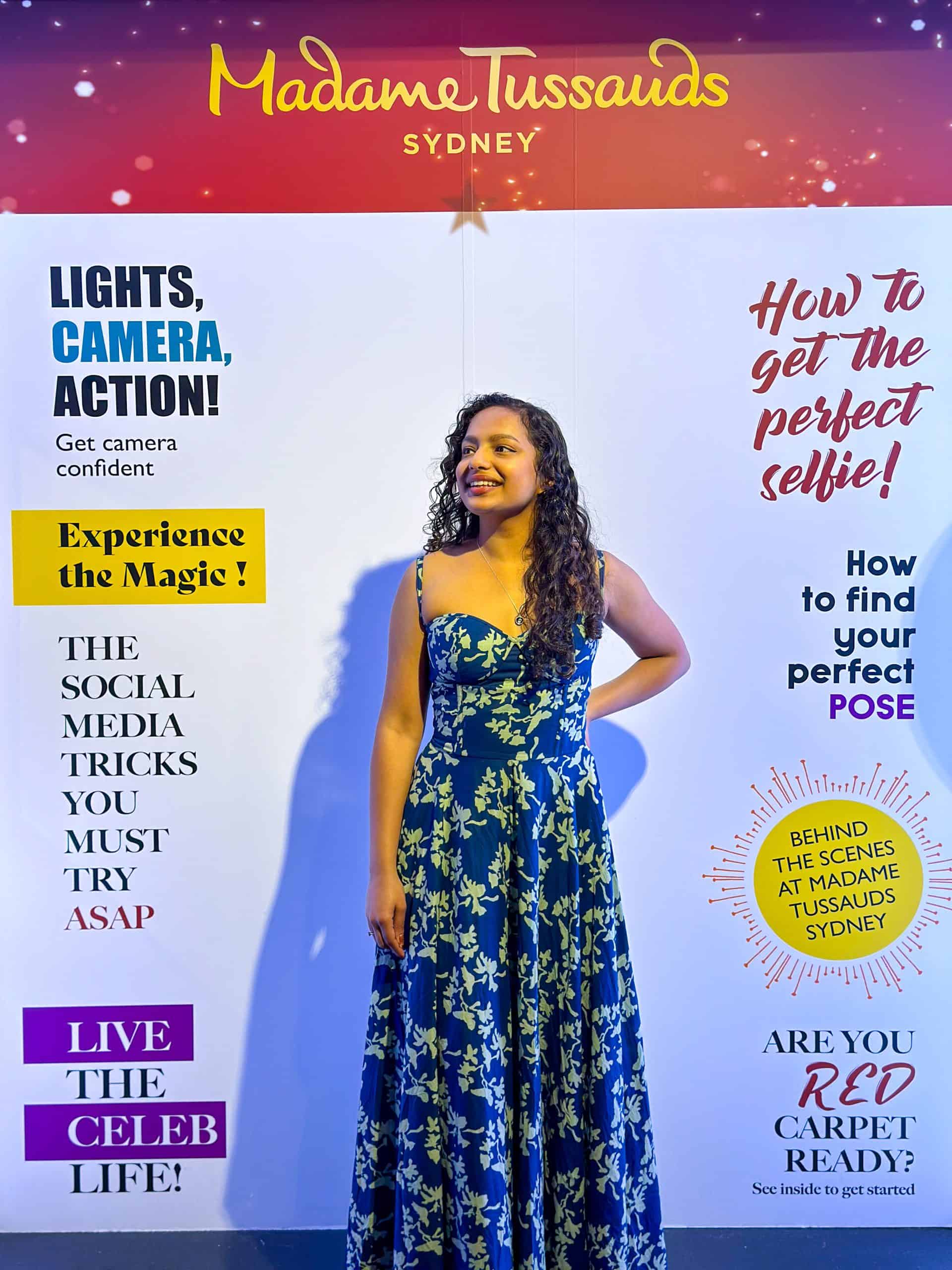 A girl wearing a blue floral dress is posing in front of a life-sized banner titled 'Madame Tussauds Sydney'.