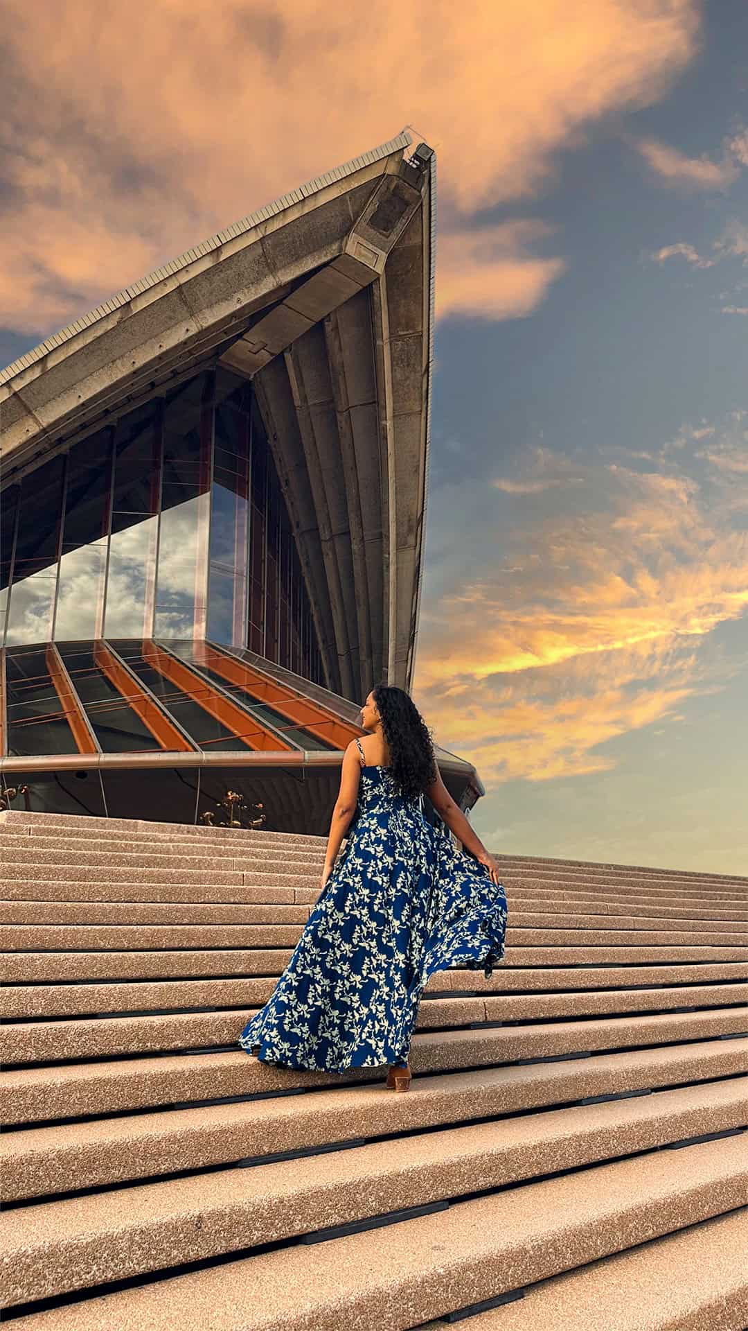 Woman in a flowing blue floral dress walks up the steps of the Sydney Opera House at golden hour.