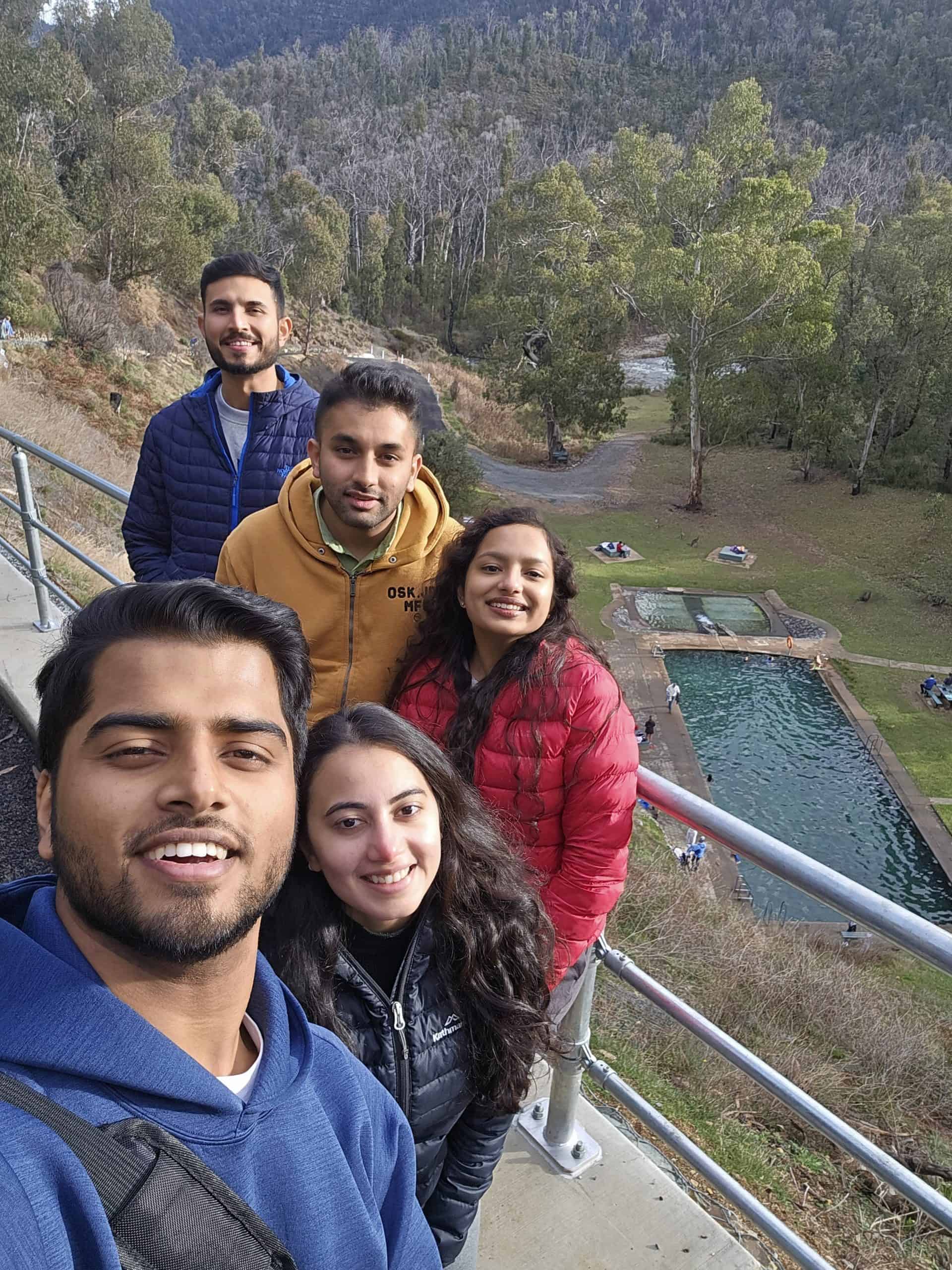 A group of friends smiles for a selfie with a scenic view of natural hot springs and lush forests.
