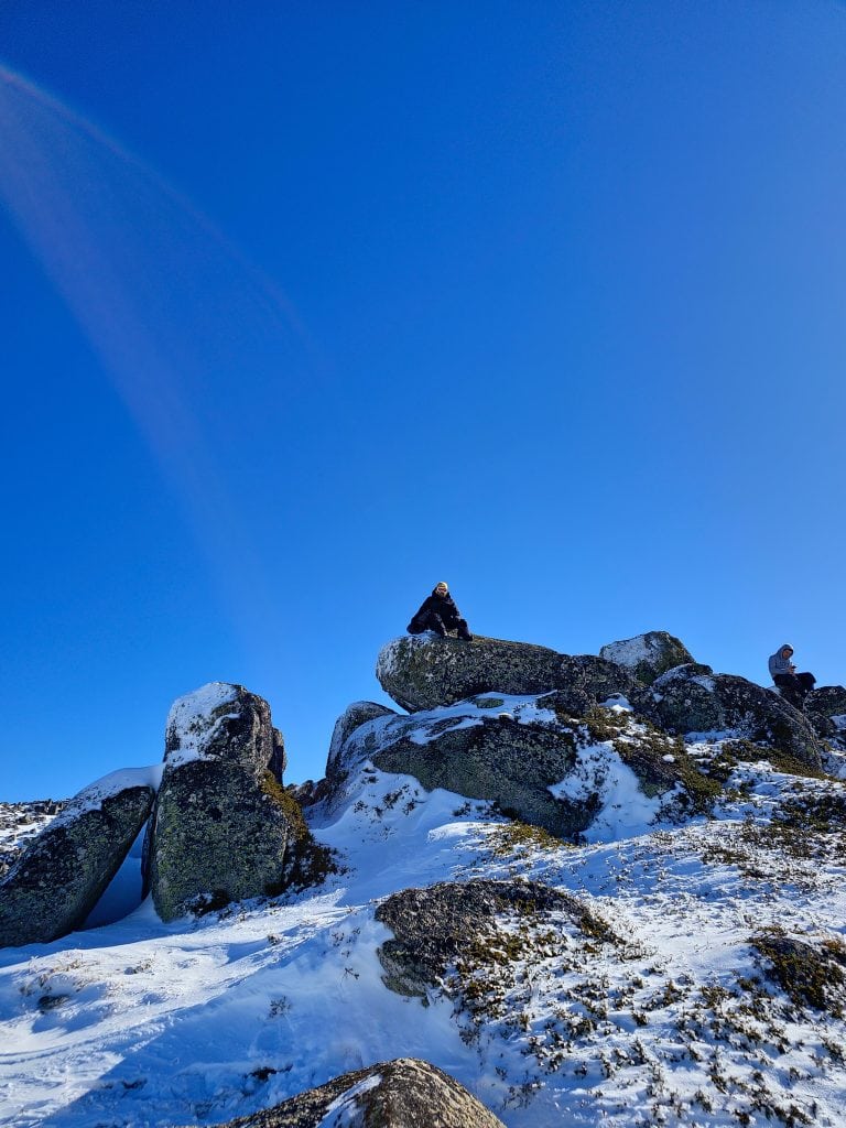 A person in winter gear sits atop large snow-dusted boulders under a bright blue sky in the Snowy Mountains, adding adventure to your snowy mountains itinerary.
