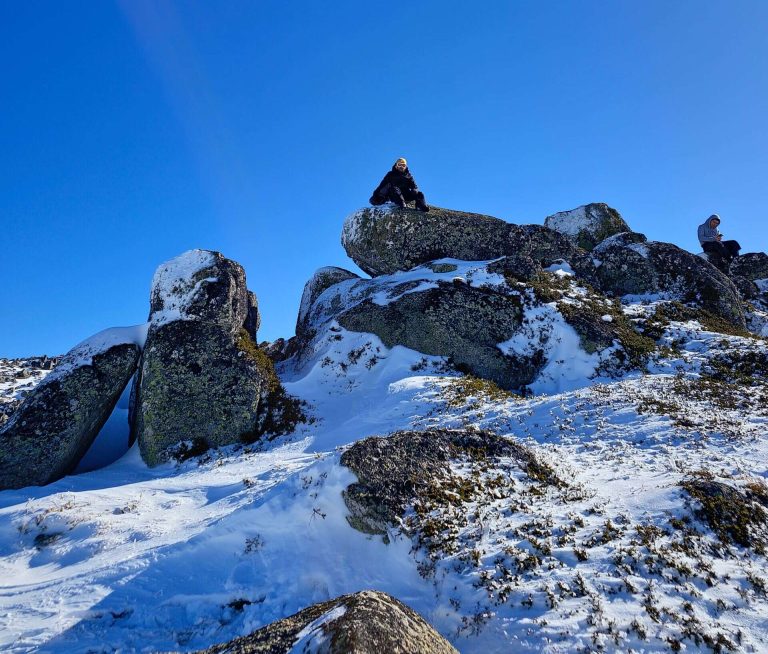 A person in winter gear sits atop large snow-dusted boulders under a bright blue sky in the Snowy Mountains, adding adventure to your snowy mountains itinerary.