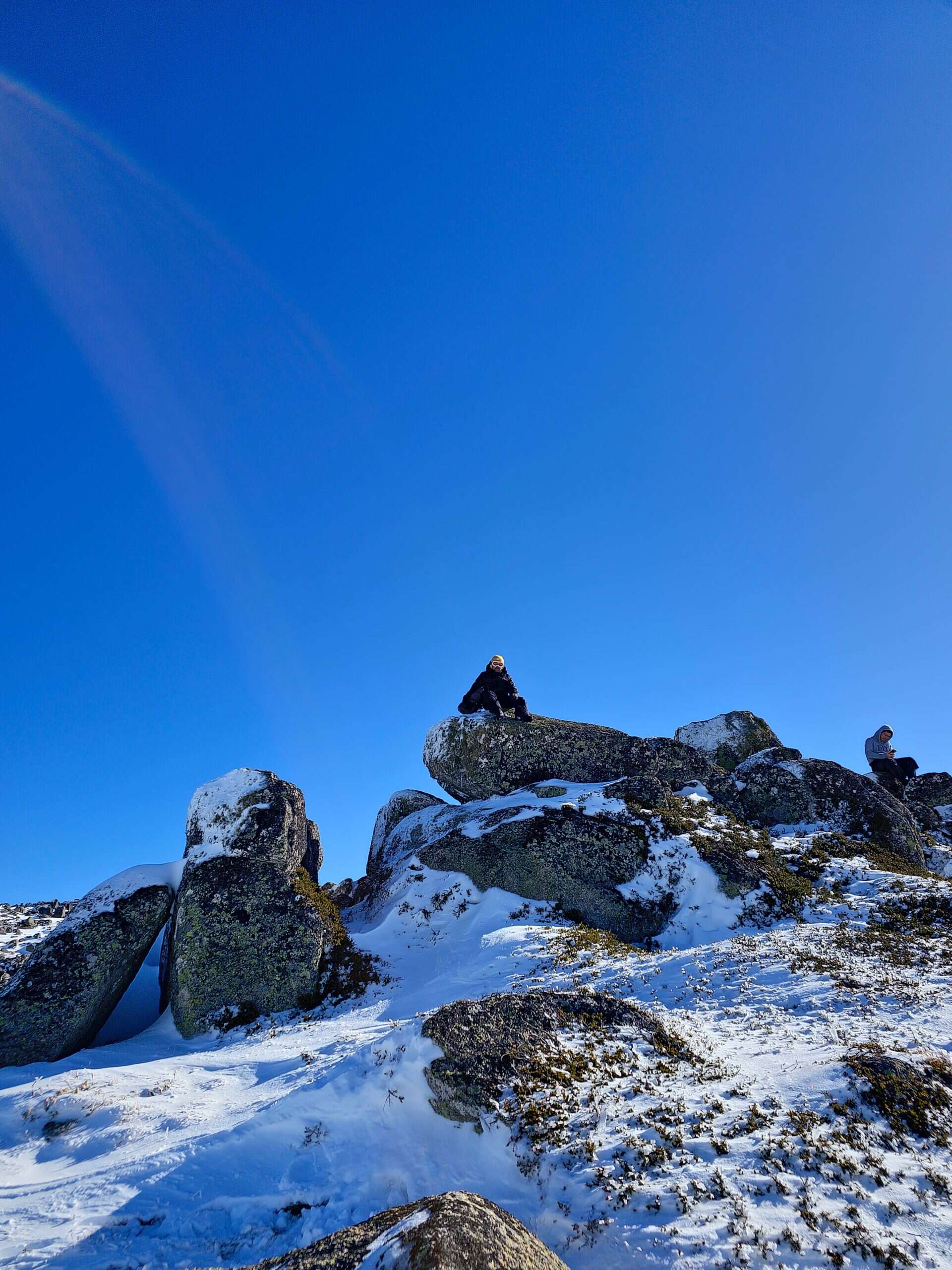 A person in winter gear sits atop large snow-dusted boulders under a bright blue sky in the Snowy Mountains, adding adventure to your snowy mountains itinerary.