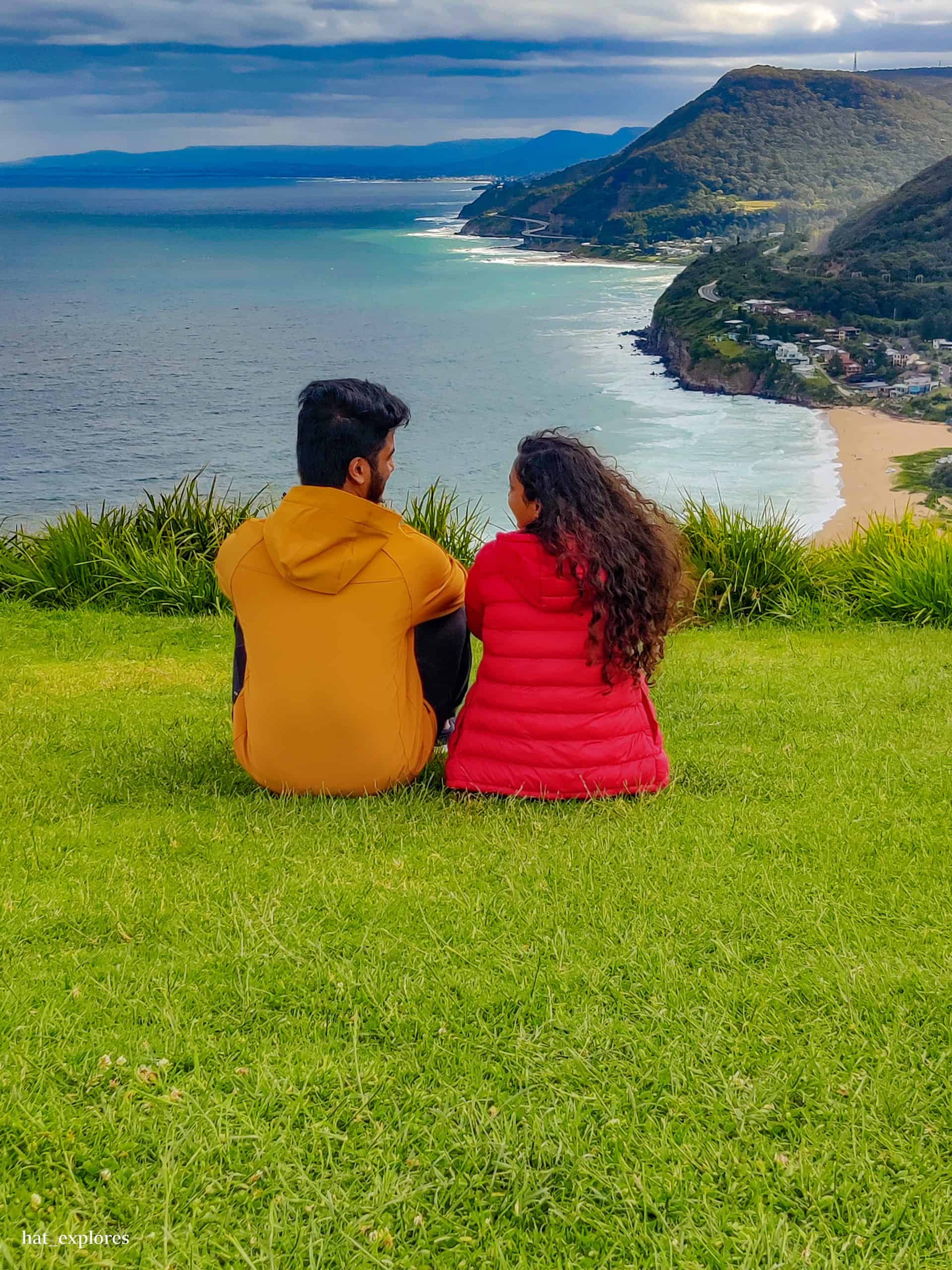 A couple sits on a grassy hilltop at Bald Hill Lookout, gazing over the coastline and Sea Cliff Bridge.