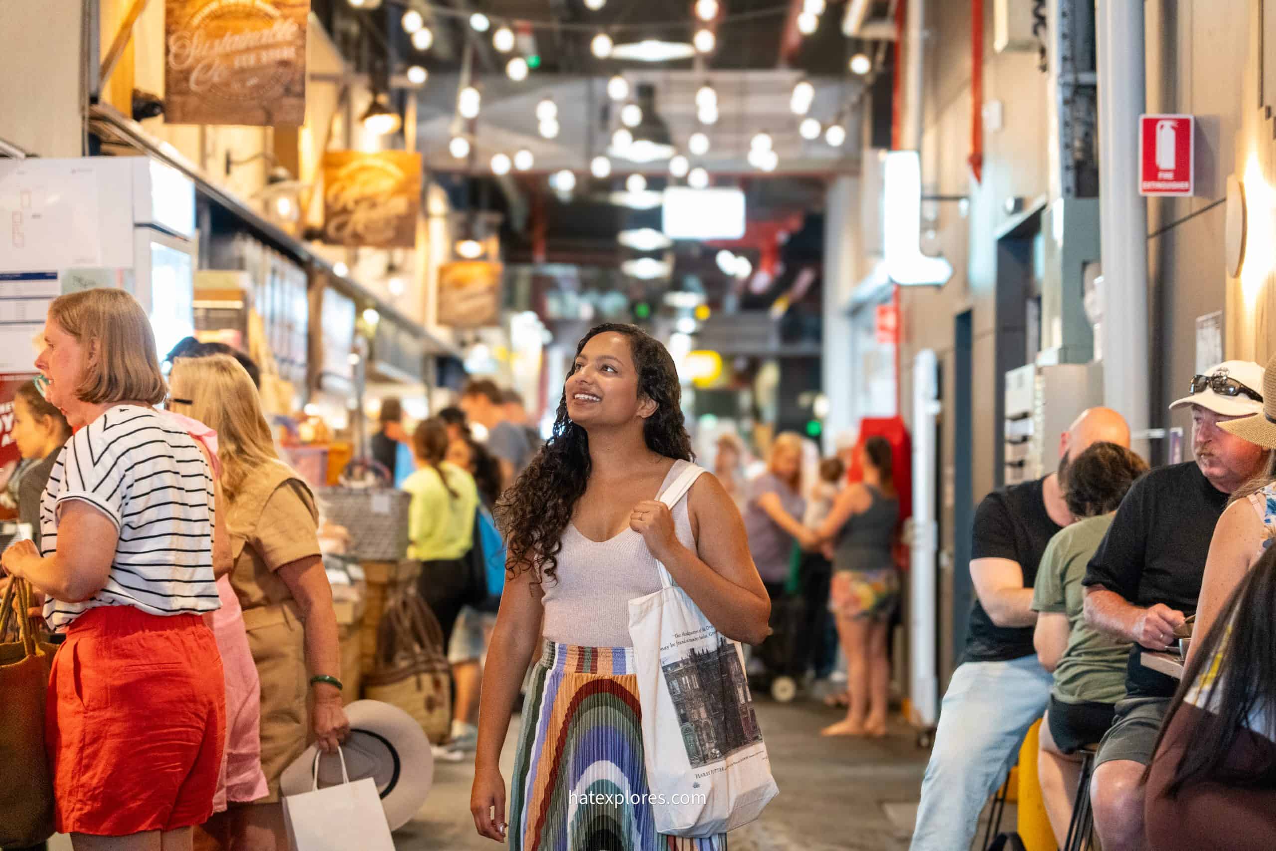 A smiling woman with a tote bag admires the lively atmosphere of Adelaide Central Market, a vibrant destination to start your culinary journey on a 3 day Adelaide itinerary.