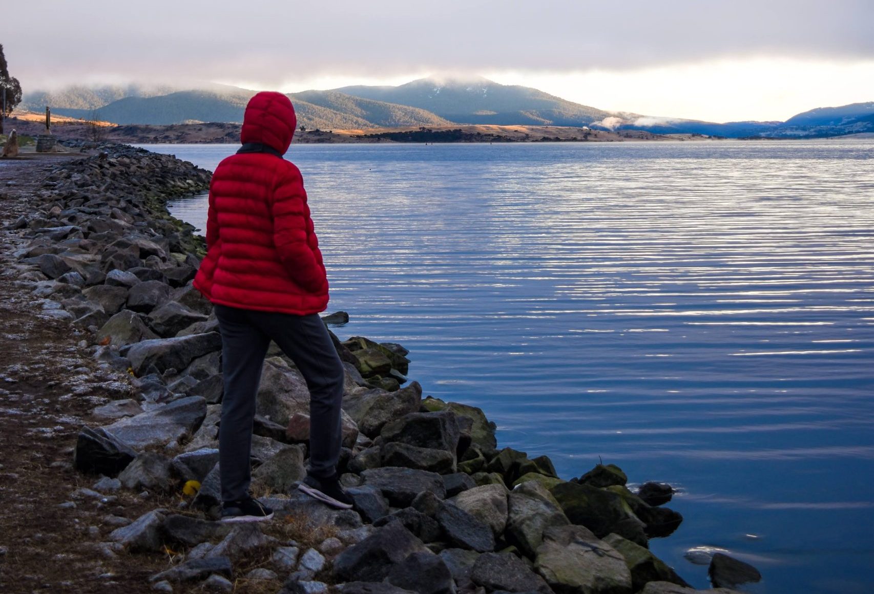 A person in a bright red jacket stands on rocky shores, gazing at a calm lake with Snowy Mountains in the distance—capturing the peaceful scenery you encounter on your road trip when figuring out how to get to Snowy Mountains from Sydney.