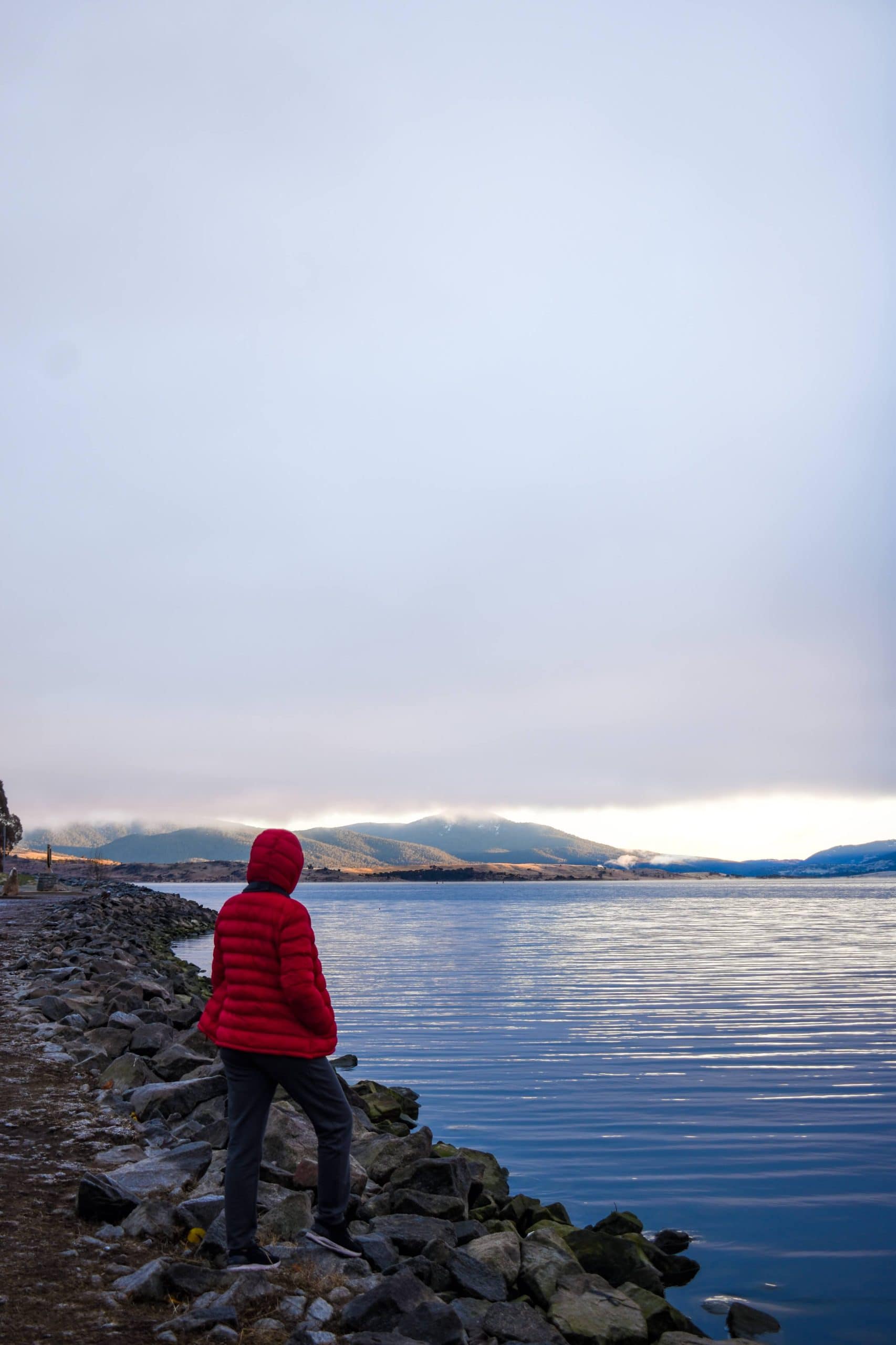 A person in a bright red jacket stands on rocky shores, gazing at a calm lake with Snowy Mountains in the distance.