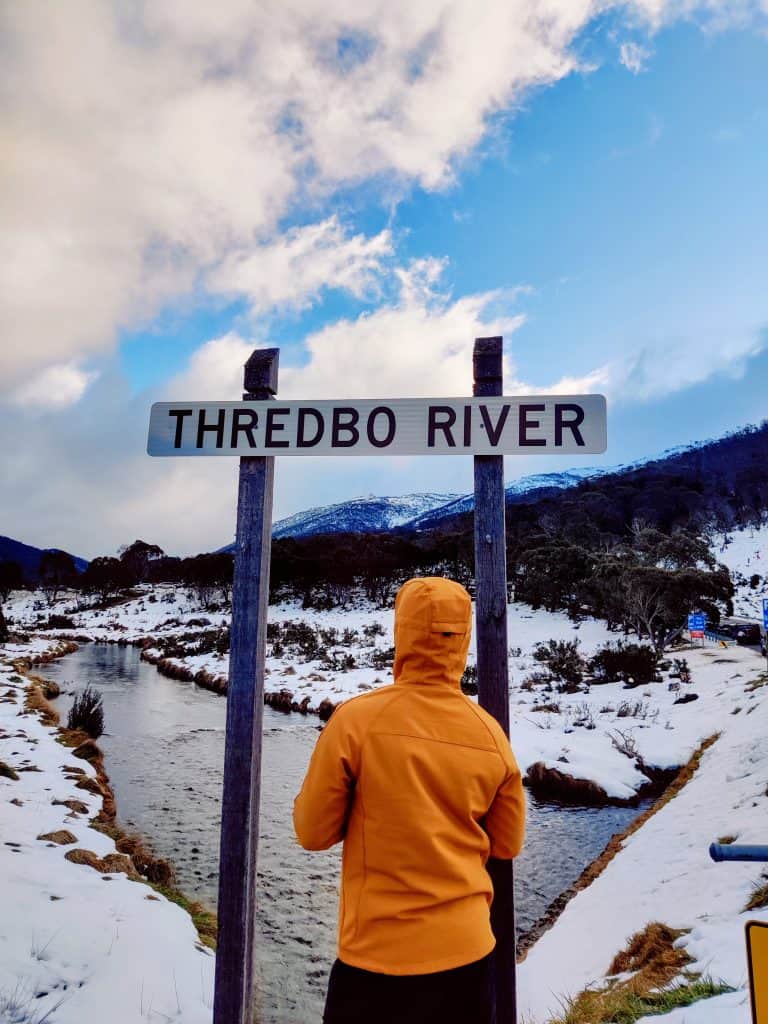 A traveler in an orange jacket stands under the Thredbo River sign, surrounded by snow and mountains, marking an iconic point