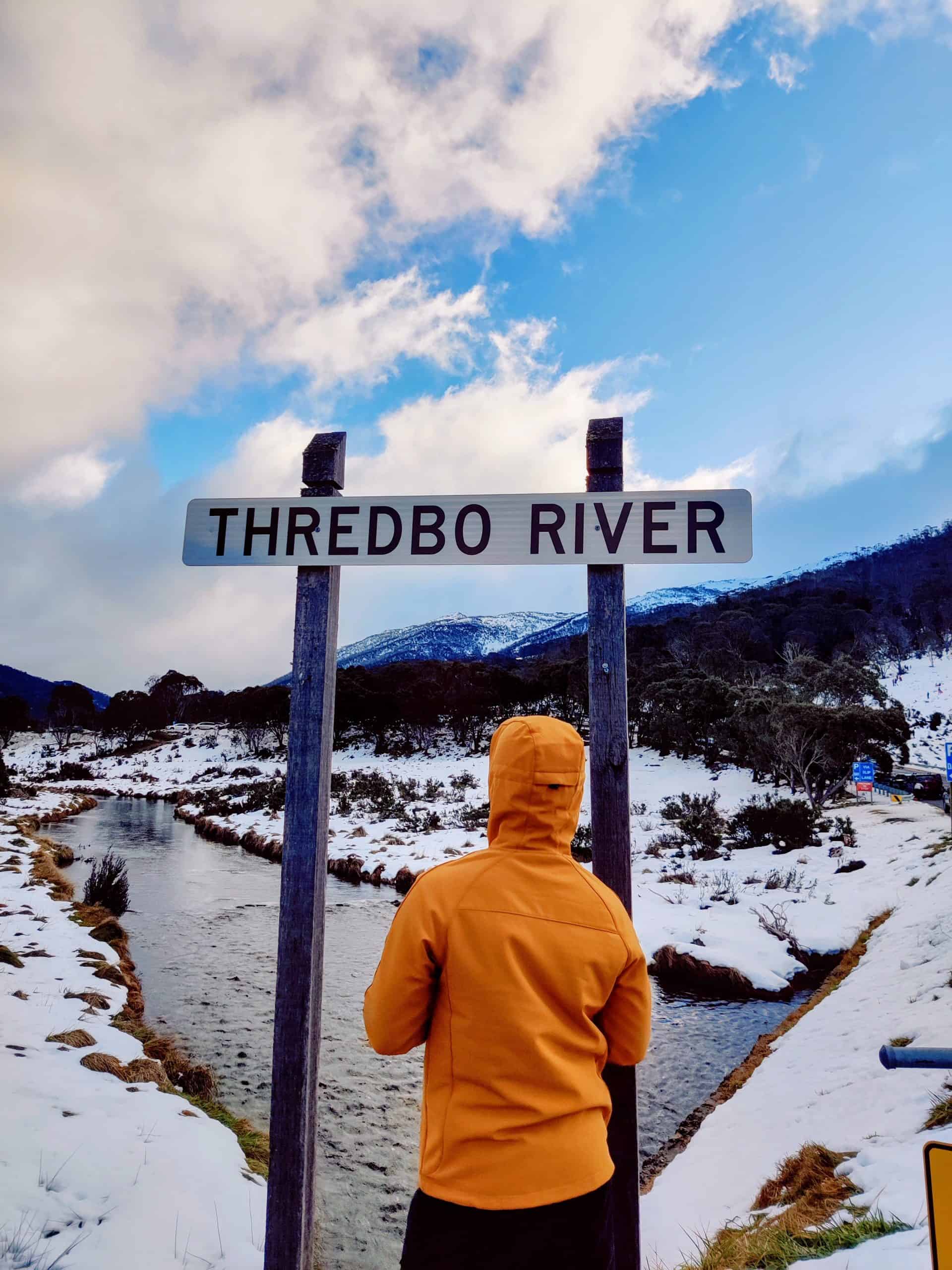 A traveler in an orange jacket stands under the Thredbo River sign, surrounded by snow and mountains, marking an iconic point.
