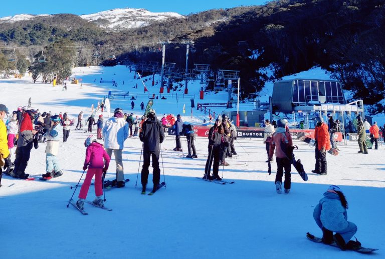 Crowds gather at the base of a snowy ski slope in the Snowy Mountains, with families and skiers preparing for their day.