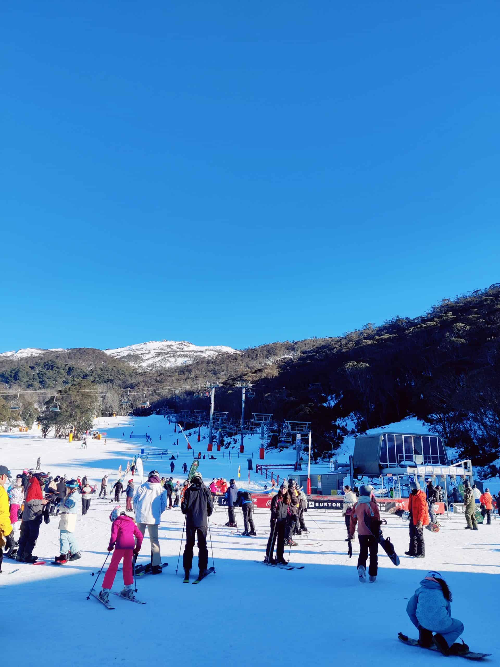 Crowds gather at the base of a snowy ski slope in the Snowy Mountains, with families and skiers preparing for their day.