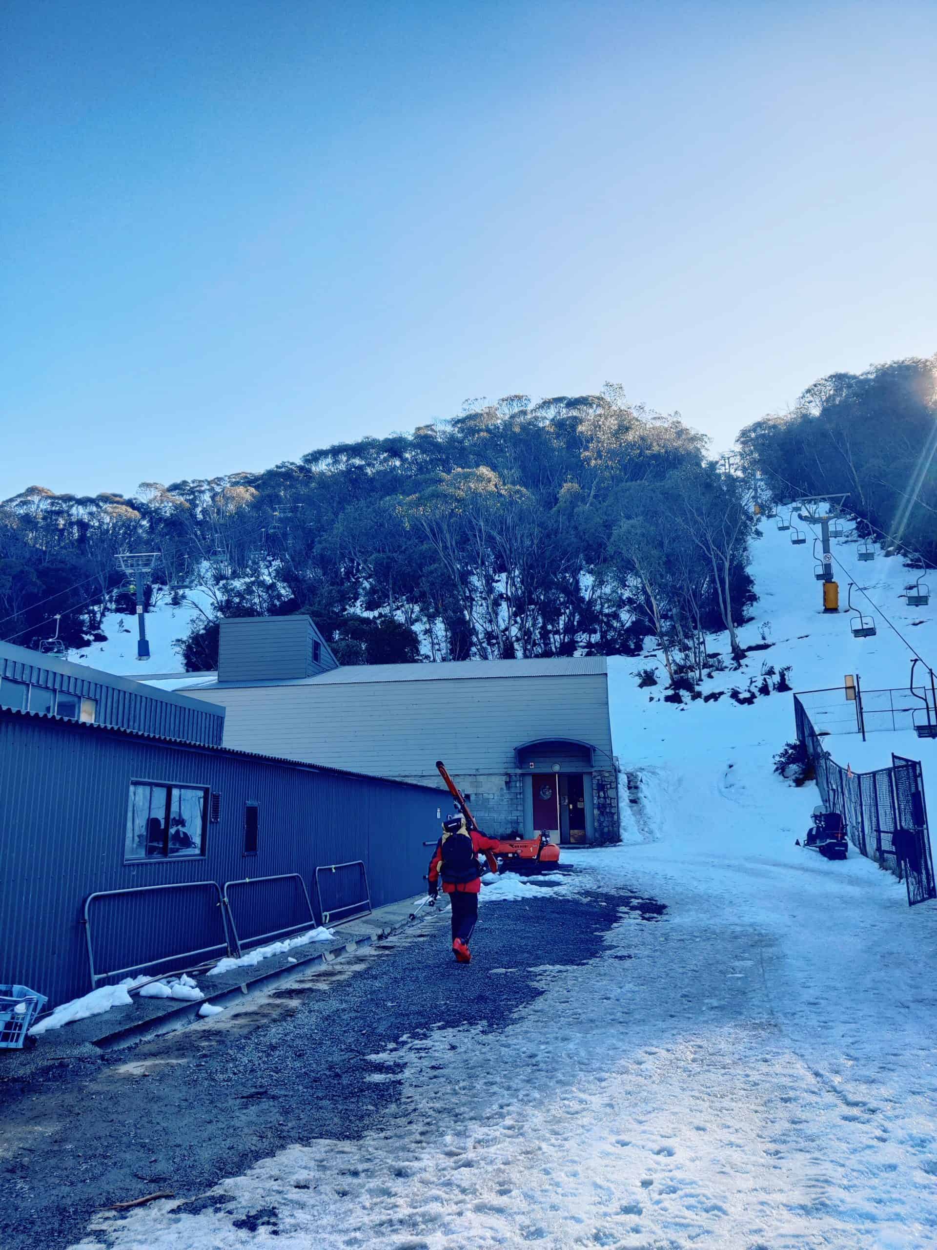 A skier carries equipment uphill past cabins and ski lifts in the Snowy Mountains,