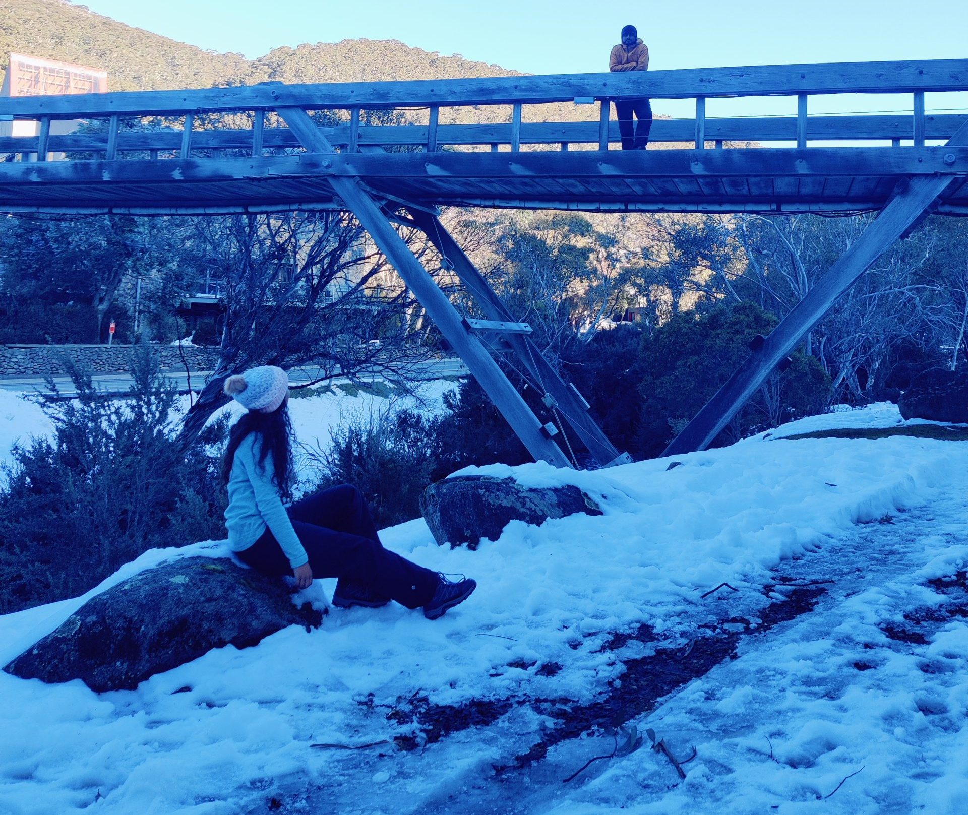 A woman sits on snowy rocks while another person stands on a rustic wooden bridge in the Snowy Mountains, illustrating picturesque stops and scenes you’ll experience while learning how to get to Snowy Mountains from Sydney.