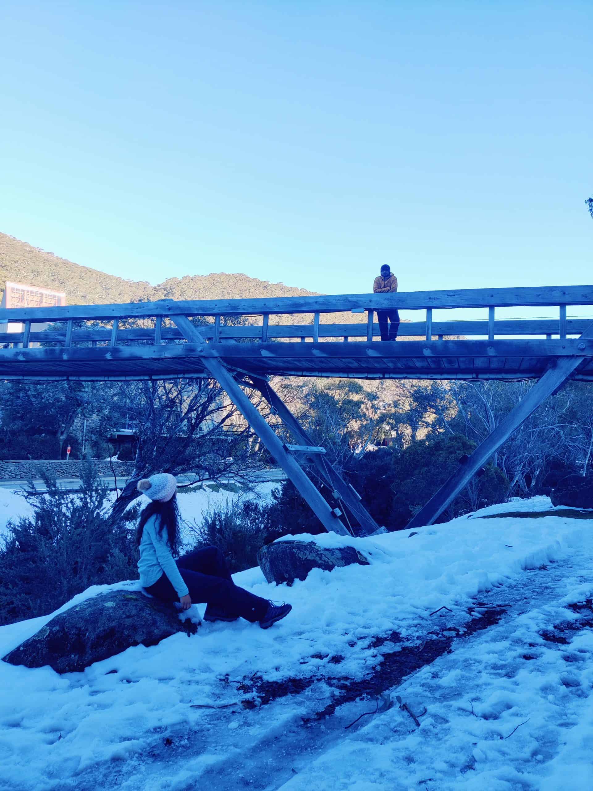 A woman sits on snowy rocks while another person stands on a rustic wooden bridge in the Snowy Mountains, illustrating picturesque stops and scenes you’ll experience while learning how to get to Snowy Mountains from Sydney.