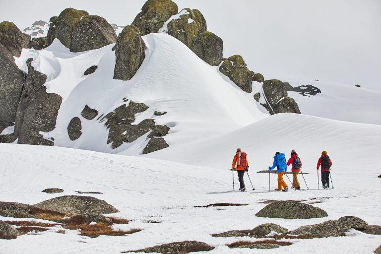 A group of four hikers in bright winter gear trek through a snow-covered landscape in the Snowy Mountains, with dramatic rocky outcrops and smooth white slopes surrounding them, perfect for a scenic and adventurous snowy mountains itinerary.