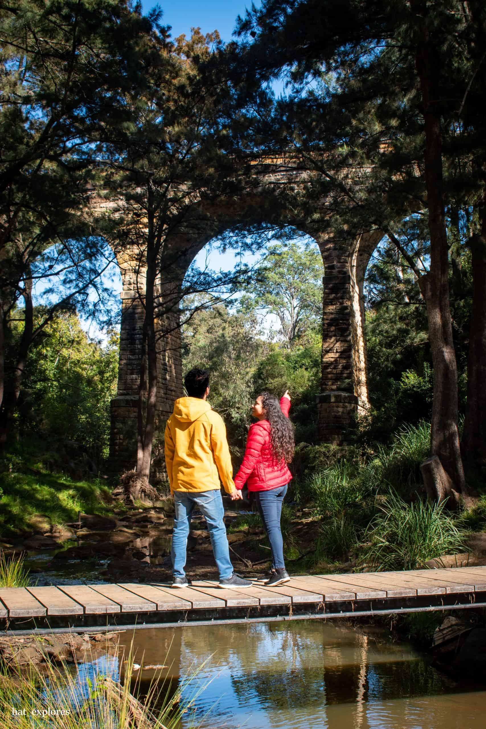 Two people hold hands on a small wooden bridge beneath the historic Picton Railway Viaduct in Wollondilly, surrounded by tall trees.