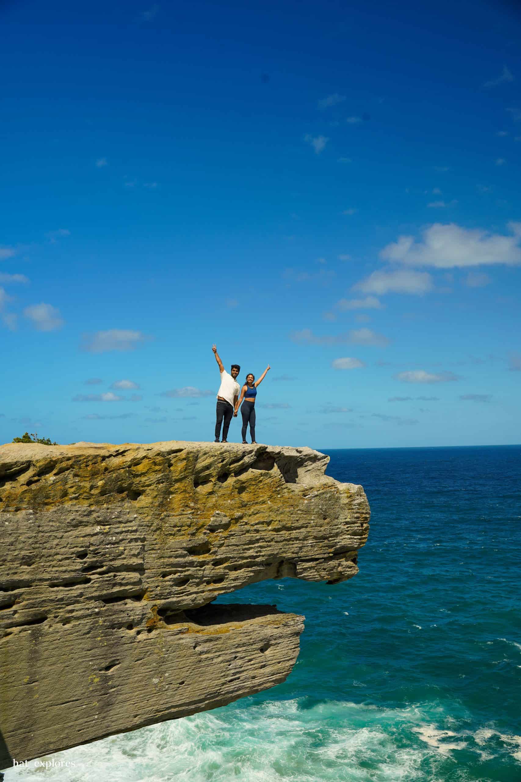 A couple stands triumphantly on the edge of Eagle Rock in Royal National Park, with the vast blue ocean stretching behind them—an iconic stop on adventurous day trips from Sydney by car.