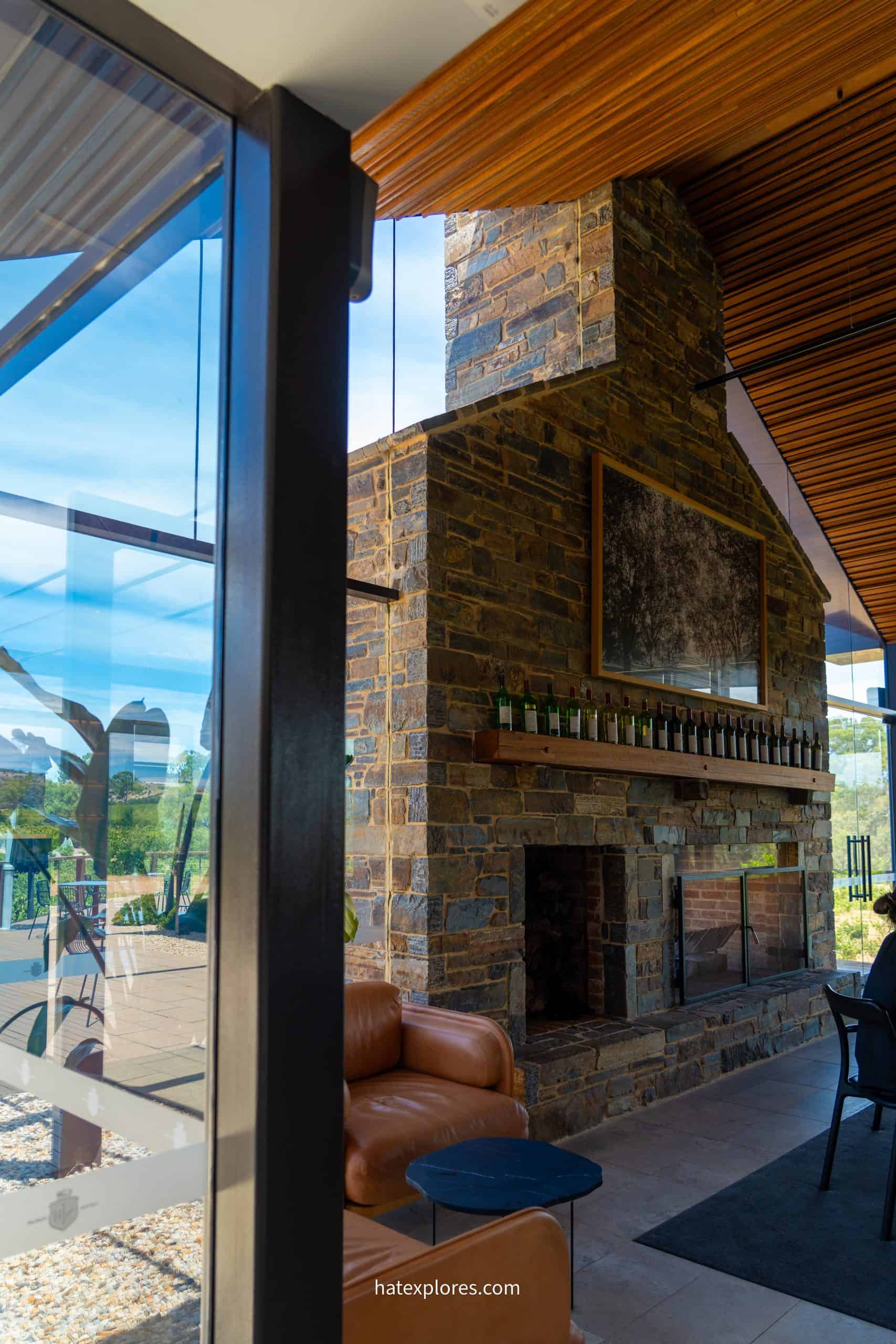 A cozy indoor lounge at a winery in Adelaide featuring a rustic stone fireplace lined with wine bottles and a large framed artwork above it, with modern glass walls overlooking a sunny vineyard. This inviting setting is a perfect wine-tasting stop on a 3-day Adelaide itinerary.