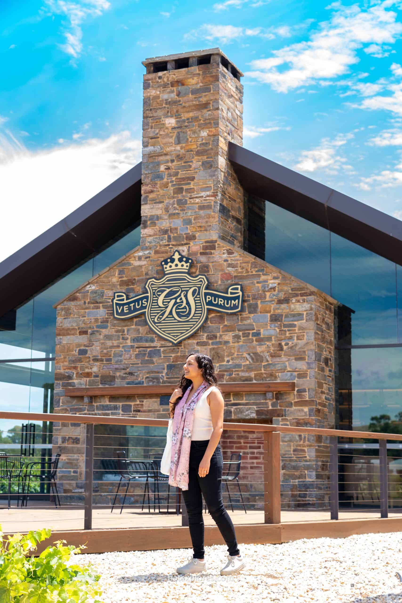  Woman standing in front of the St Hugo Wines estate in the Barossa Valley, with a modern stone and glass building behind her under a bright blue sky.