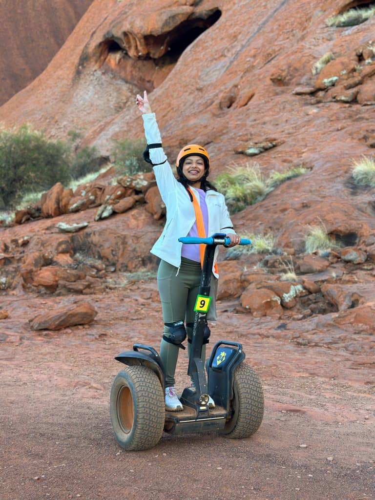 A smiling woman wearing a helmet and safety gear rides a Segway on a red dirt path at the base of Uluru, flashing a peace sign during an Uluru Segway tour. The iconic red rock formation looms in the background, creating a dramatic natural setting.