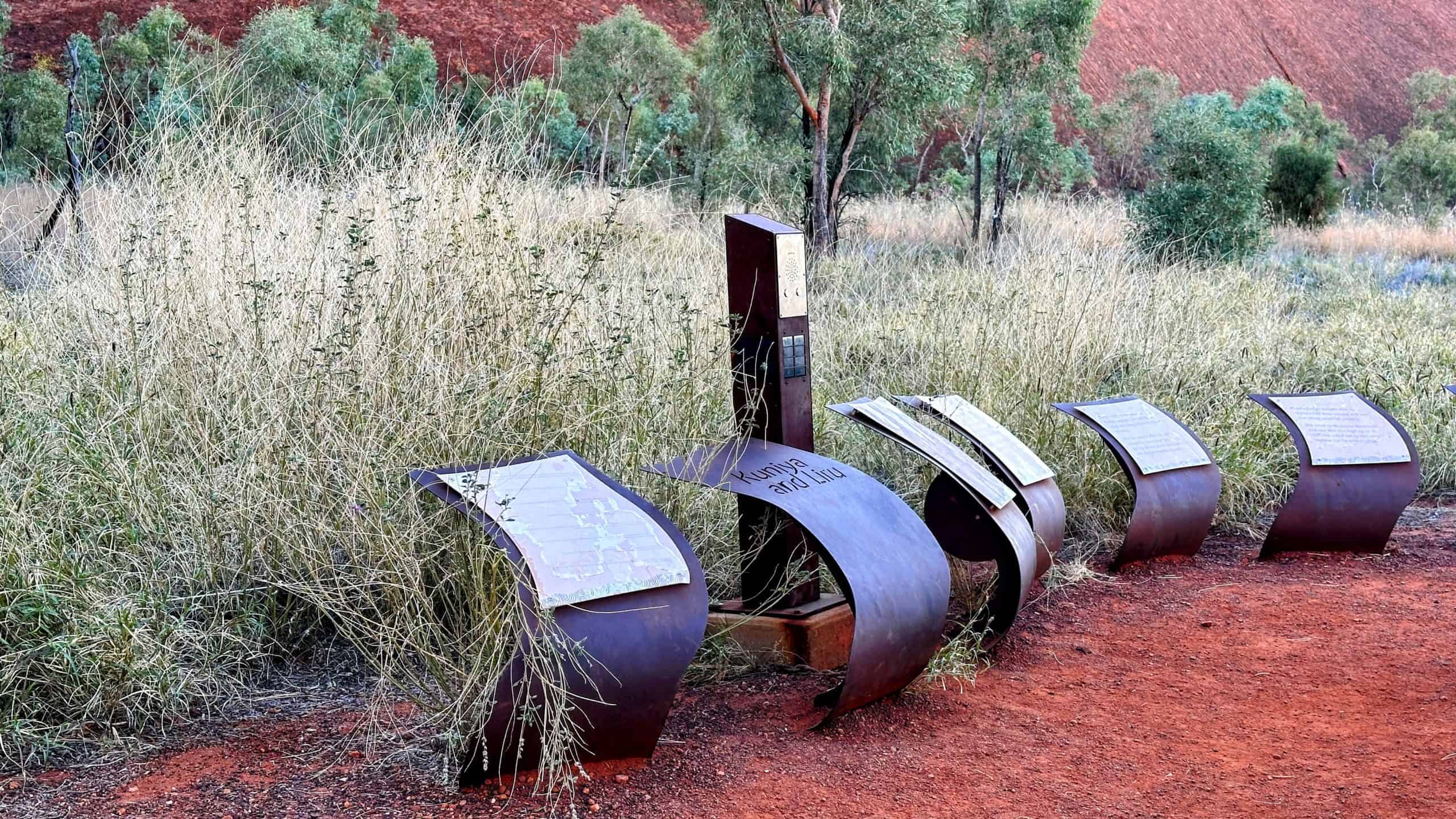 Curved metal informational signs line a trail with native grasses and trees in front of Uluru. These signs provide cultural and environmental insights into the area's Aboriginal significance.