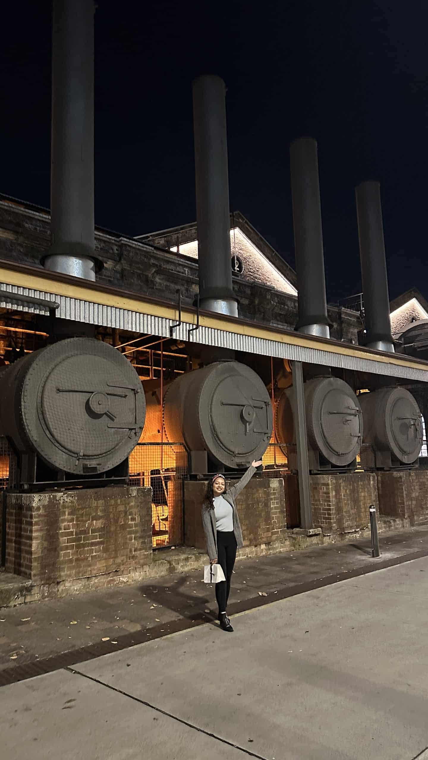 A smiling woman poses in front of a festive stall window at the Sydney winter markets, holding a brochure beside a decorative cardboard Christmas tree made of gingerbread-shaped cutouts and glowing fairy lights.