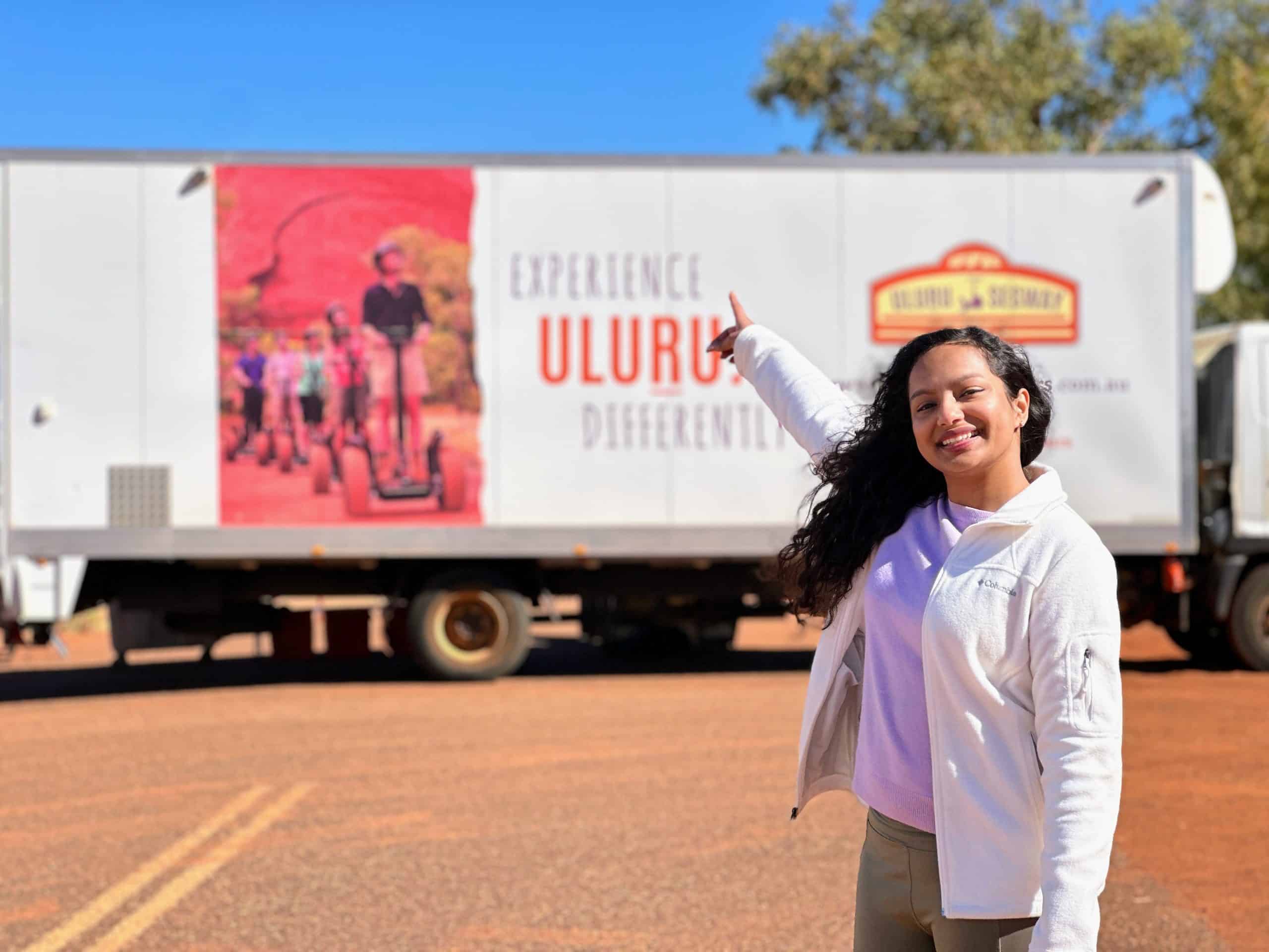 A cheerful woman points at a branded tour van promoting Uluru Segway tours, featuring a large image of a group riding Segways near the red cliffs of Uluru. The clear blue sky and outback surroundings highlight the adventure atmosphere.

